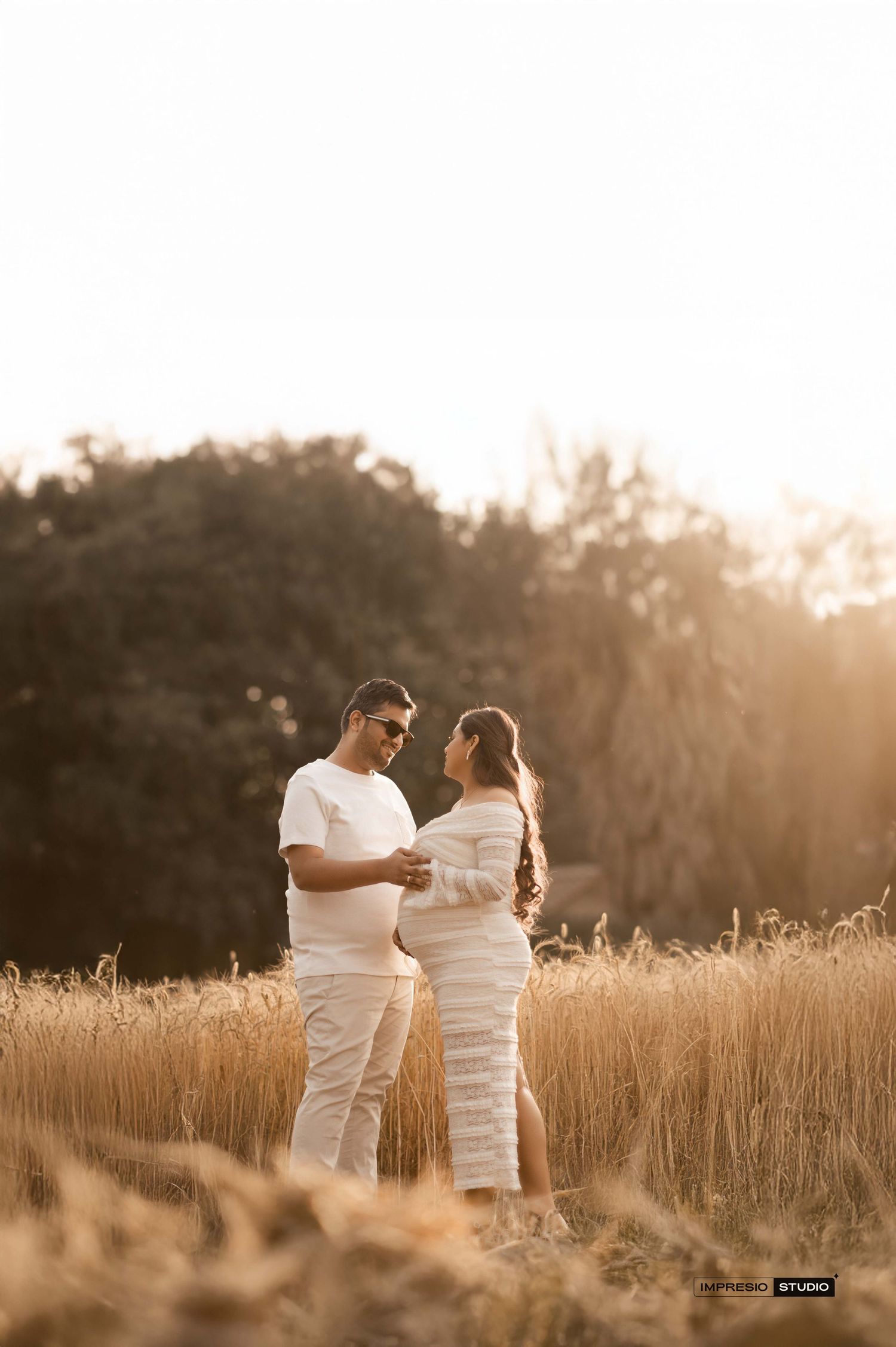Couple in white outfits sharing a tender maternity moment in a golden wheat field at sunset.