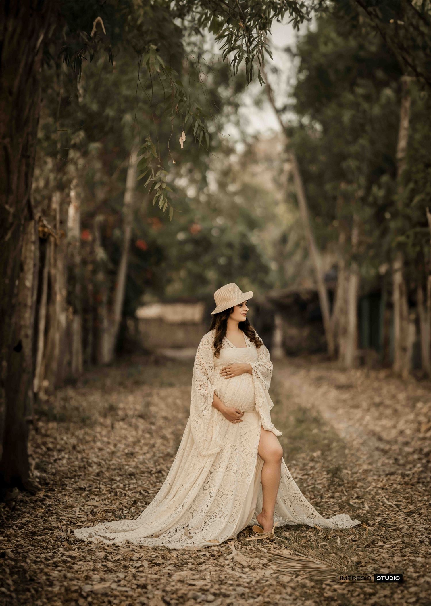 Pregnant woman in white lace gown and hat poses on a leaf-covered path surrounded by trees in a maternity photoshoot.