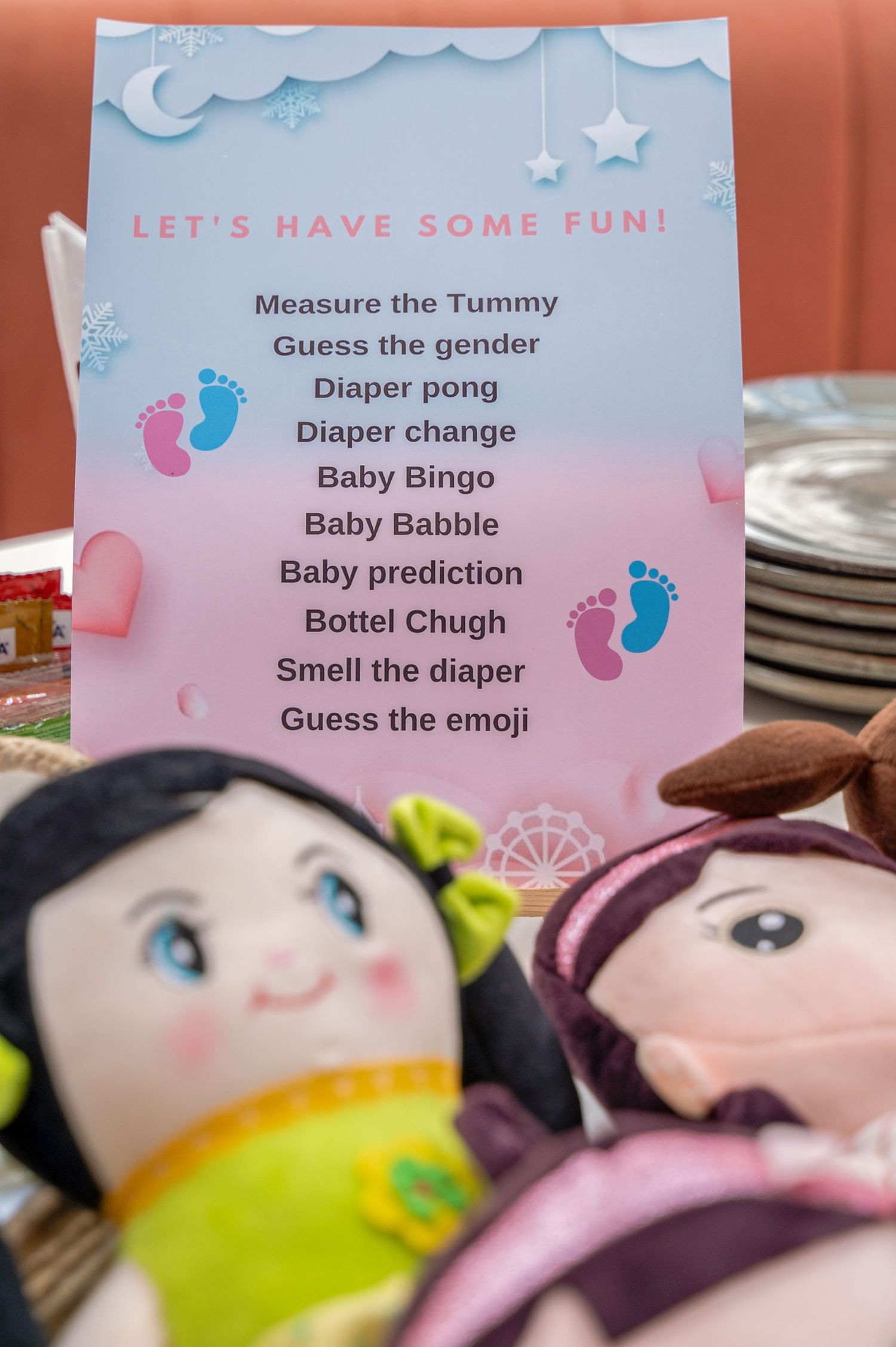 Baby shower game sign with dolls, listing fun activities like Diaper Bingo, Baby Babble, and Guess the Emoji.