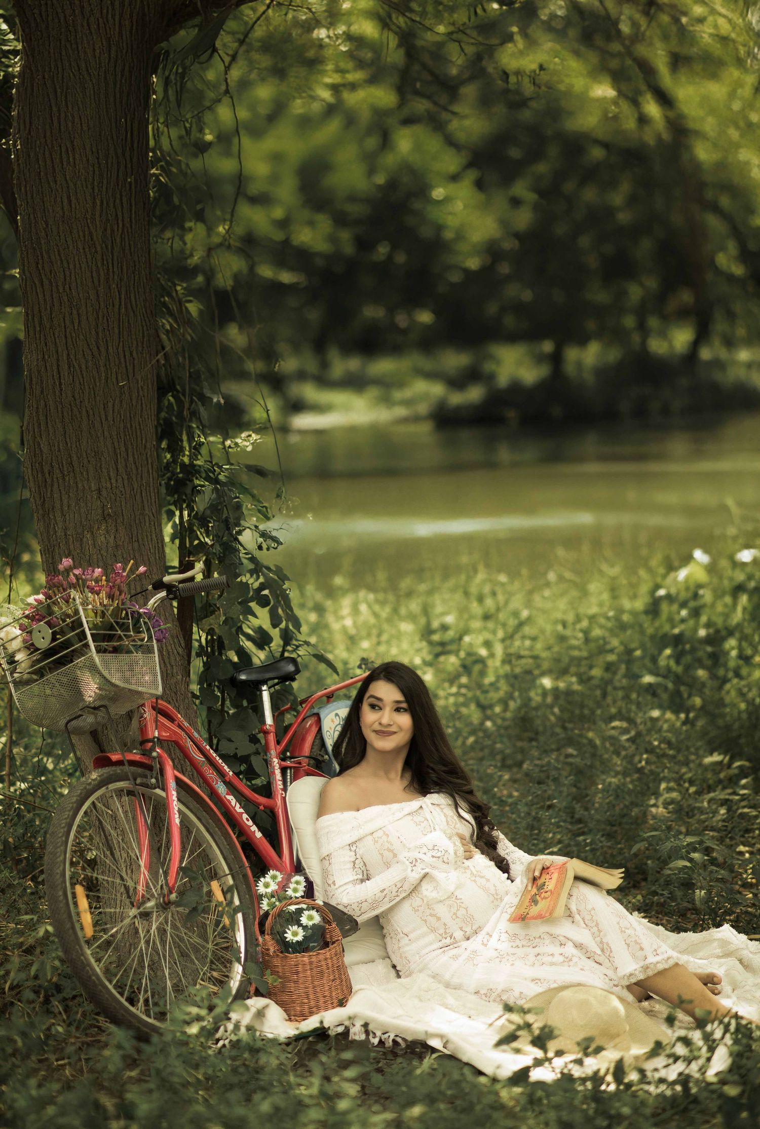 Woman in white dress reading a book by a tree near a river, with a red bicycle and picnic basket nearby.