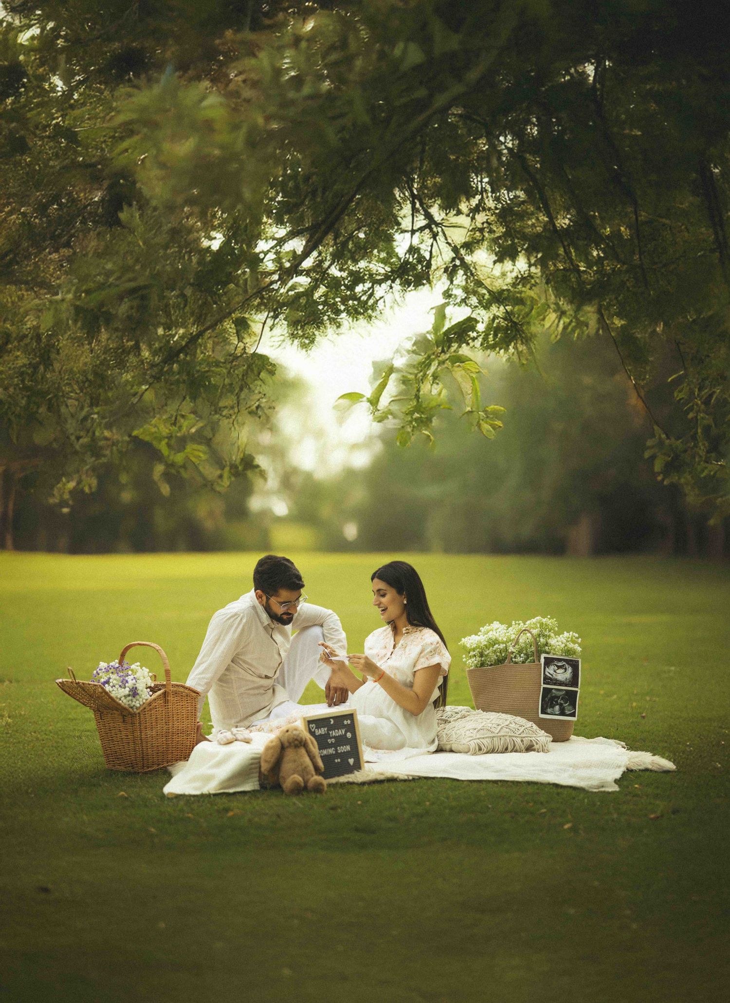 Couple sitting on white blanket in lush green park for maternity photoshoot with picnic basket and ultrasound photos.