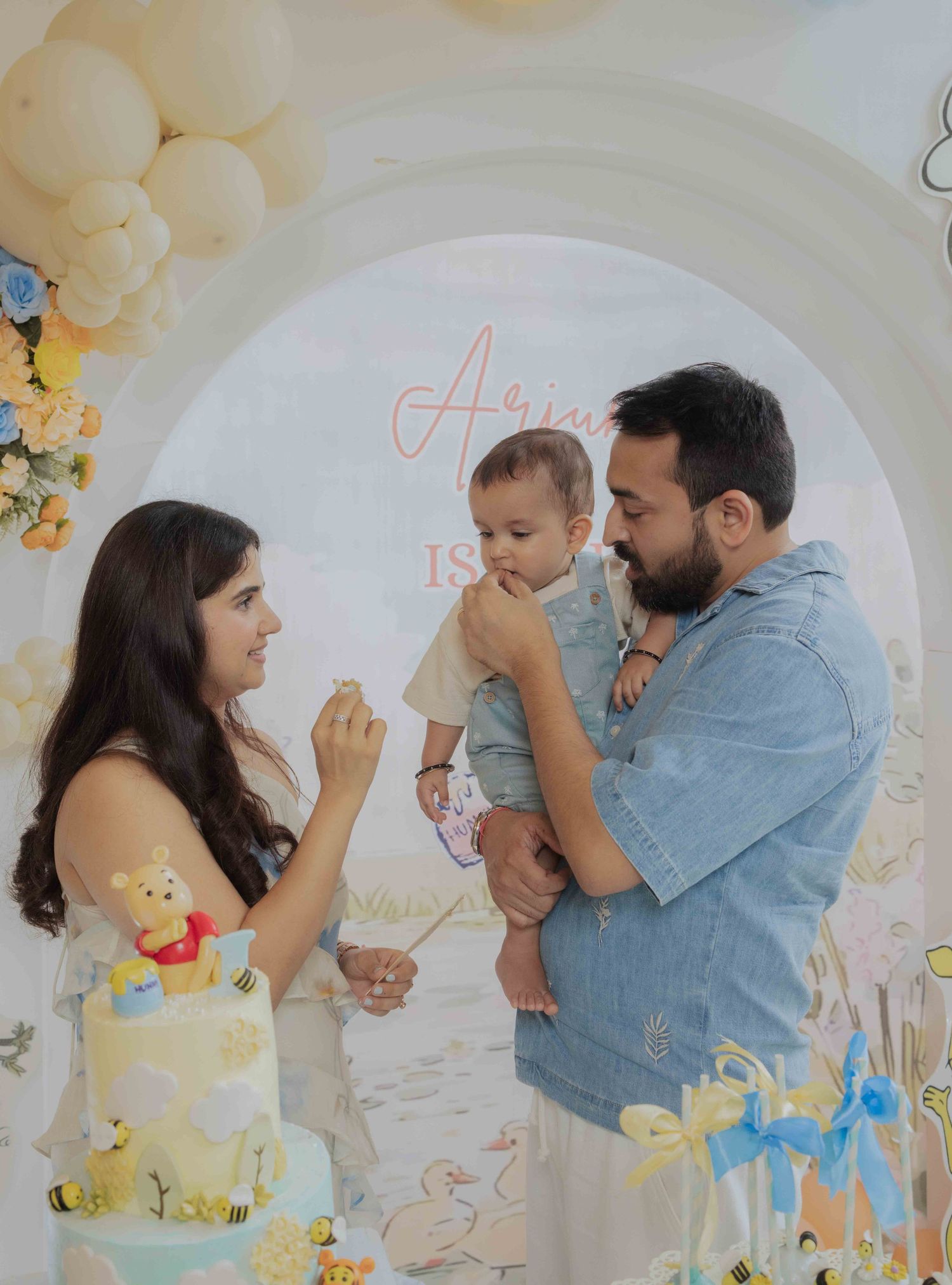 Family celebrating baby's first birthday with Winnie the Pooh themed cake and balloon arch backdrop.