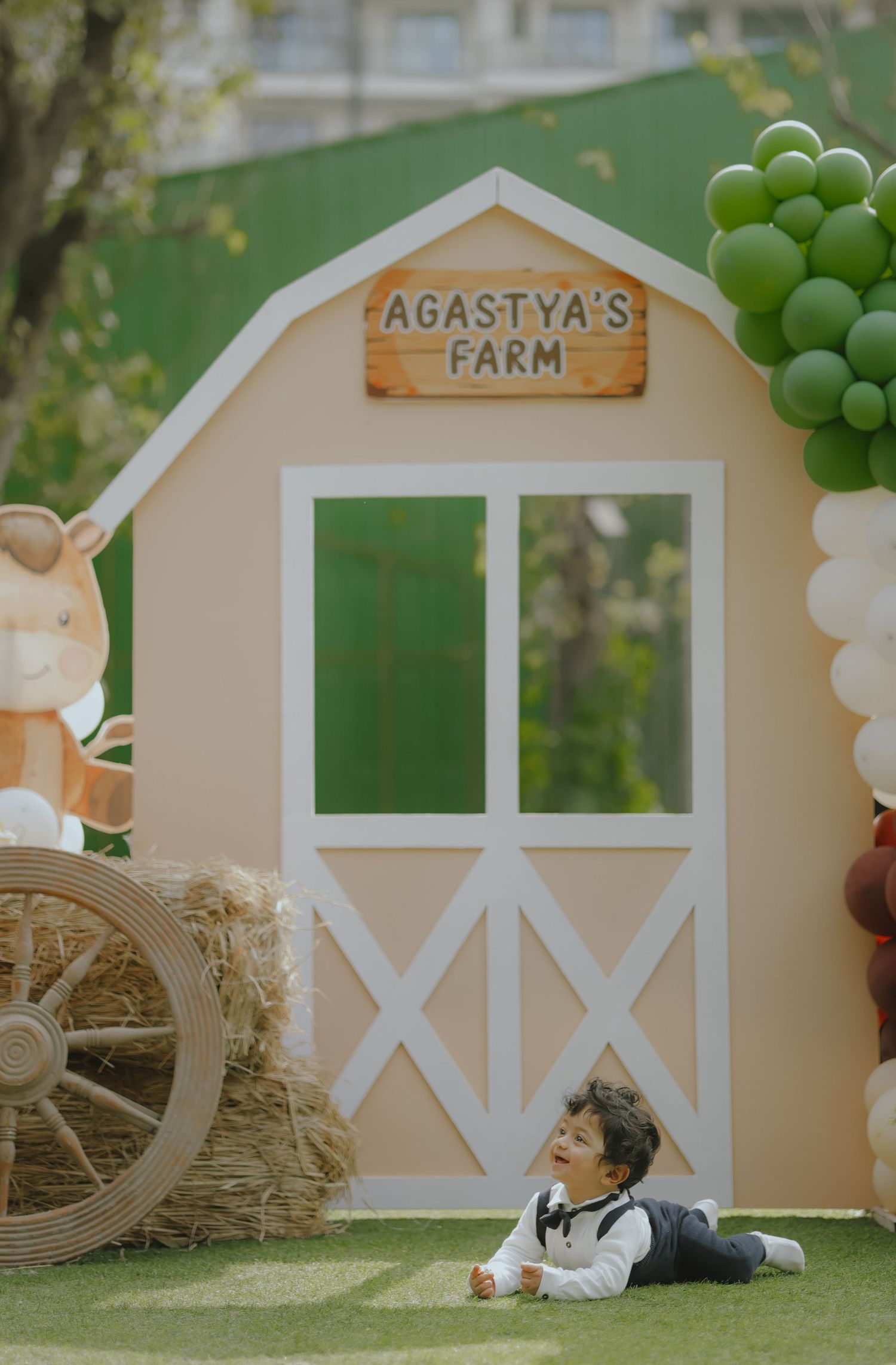 Toddler lying on grass at Agastya's Farm themed birthday party with barn backdrop and green balloon decorations.