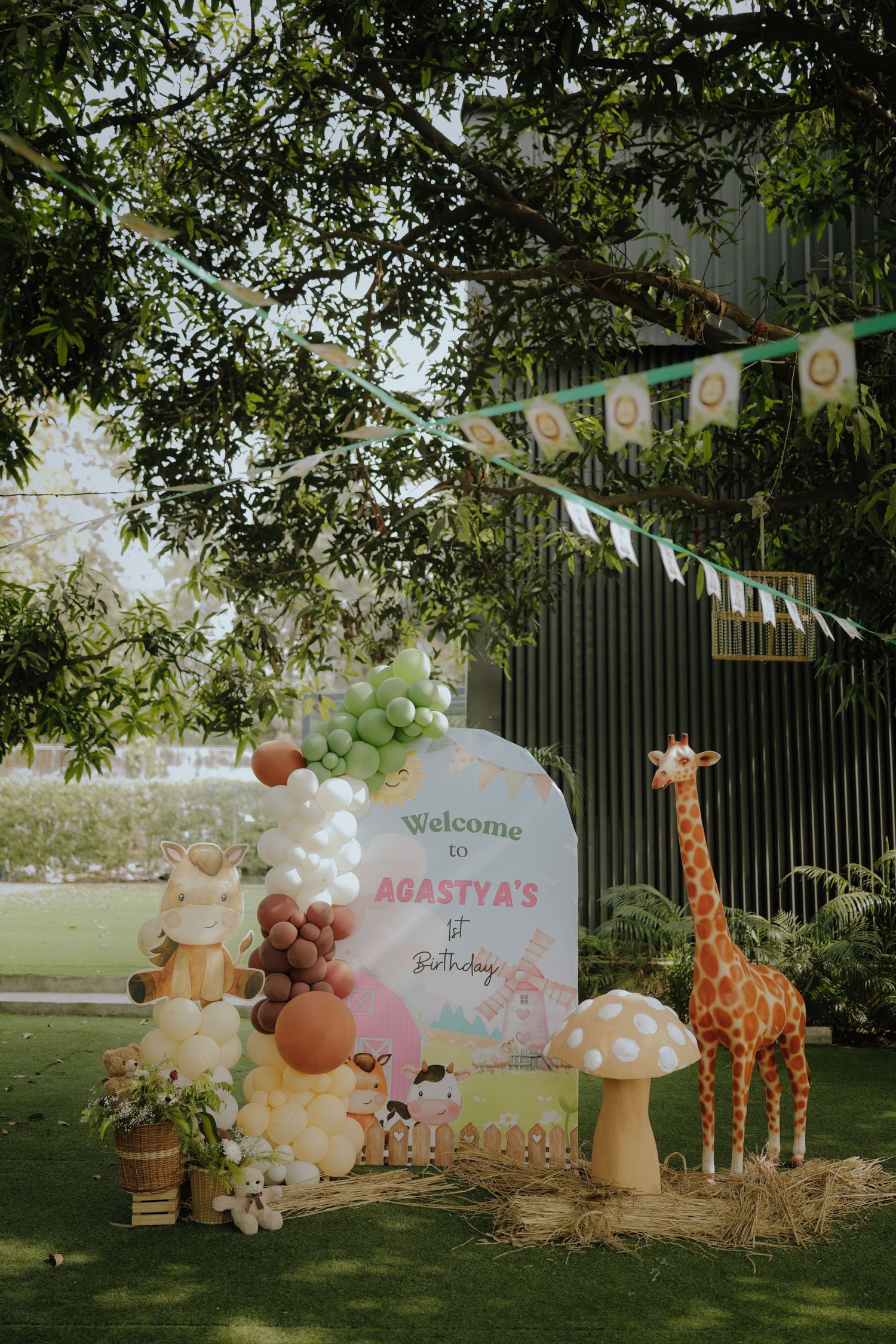Outdoor jungle safari 1st birthday party setup with balloon garland, giraffe prop, and welcome sign for Agastya.