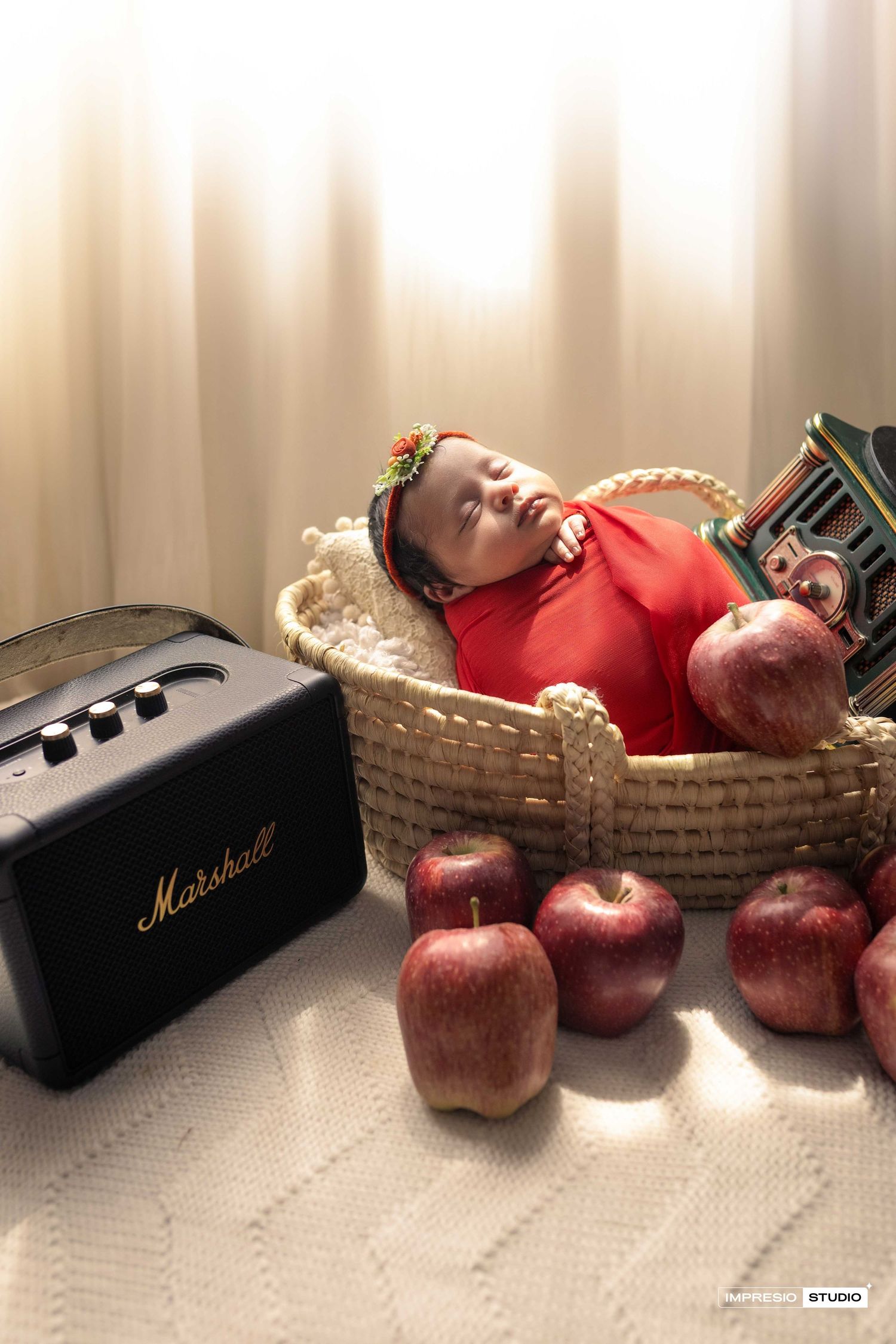 Newborn baby sleeping in wicker basket surrounded by red apples with Marshall speaker beside, soft studio lighting.
