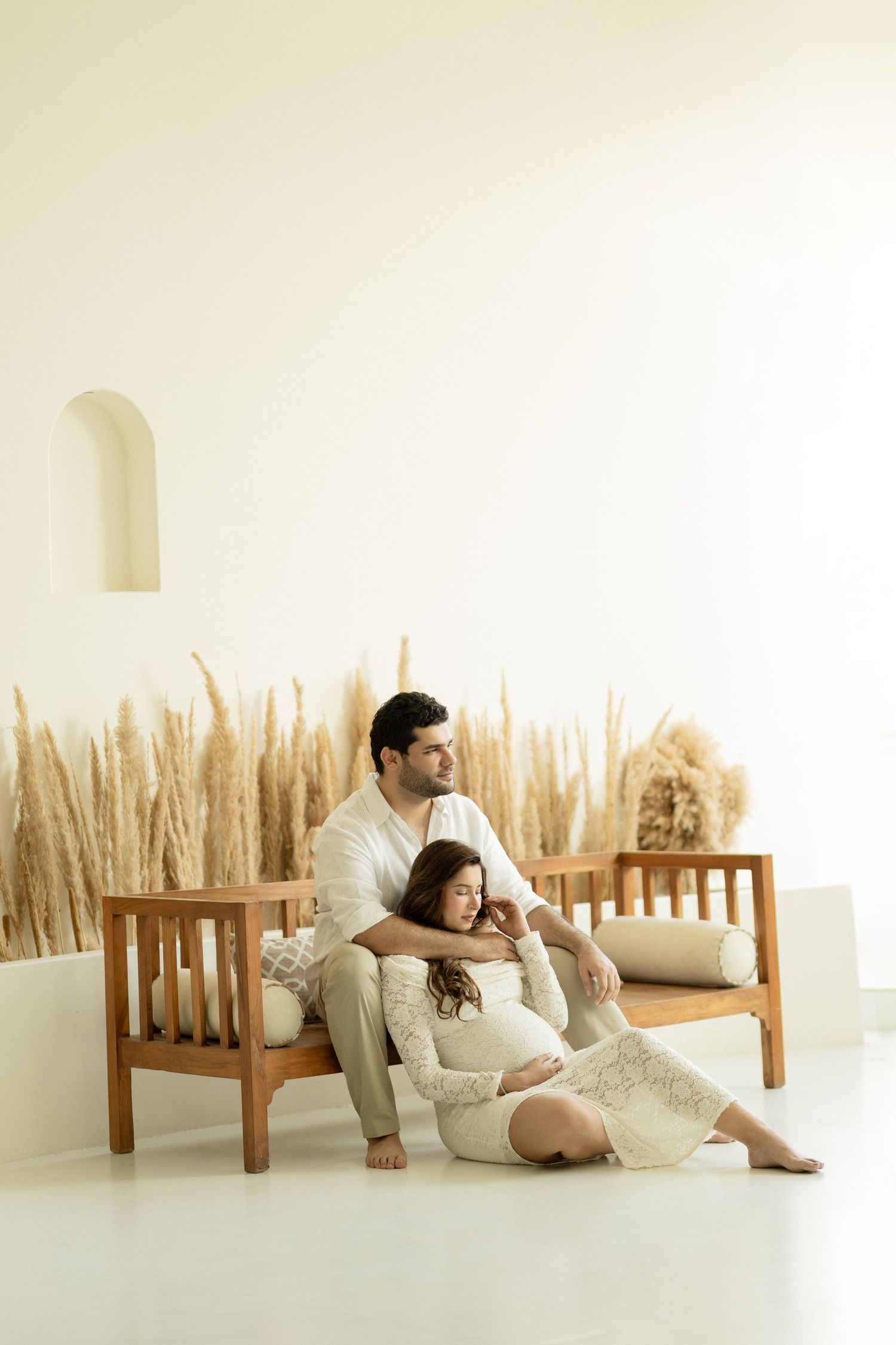 Couple in white clothing relaxing on wooden furniture in a minimalist white studio setting with natural decor.
