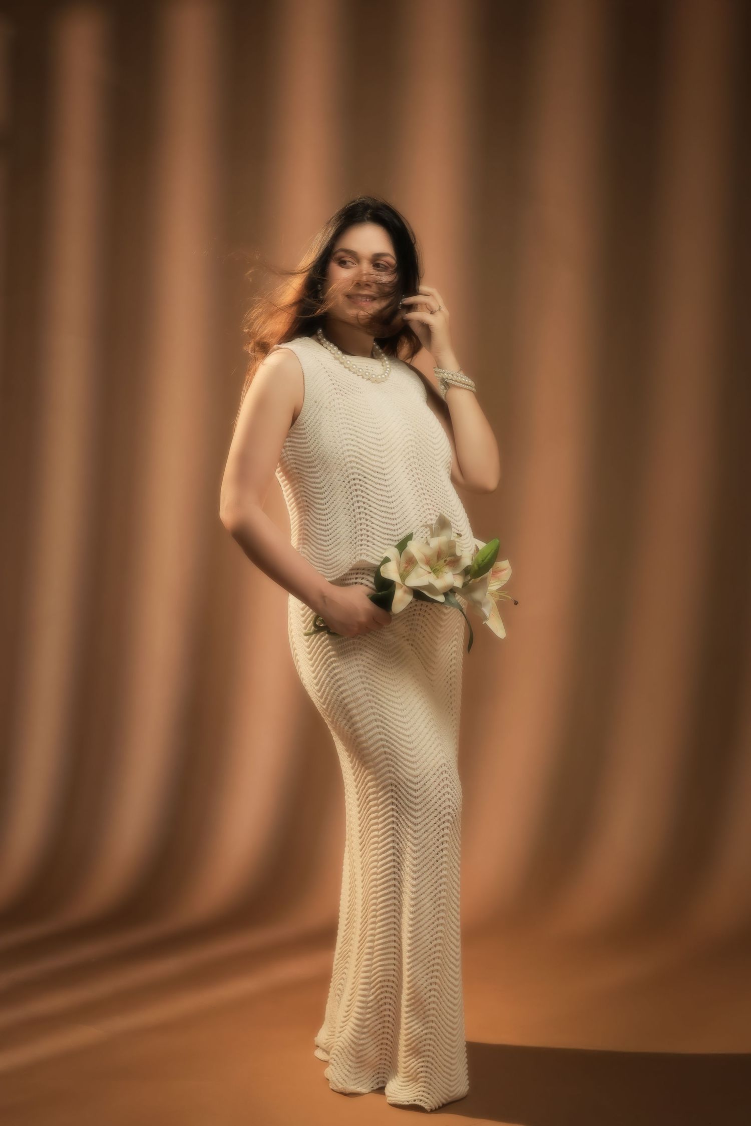 A person in a cream-colored knit dress holds white flowers while posing against a brown draped backdrop.