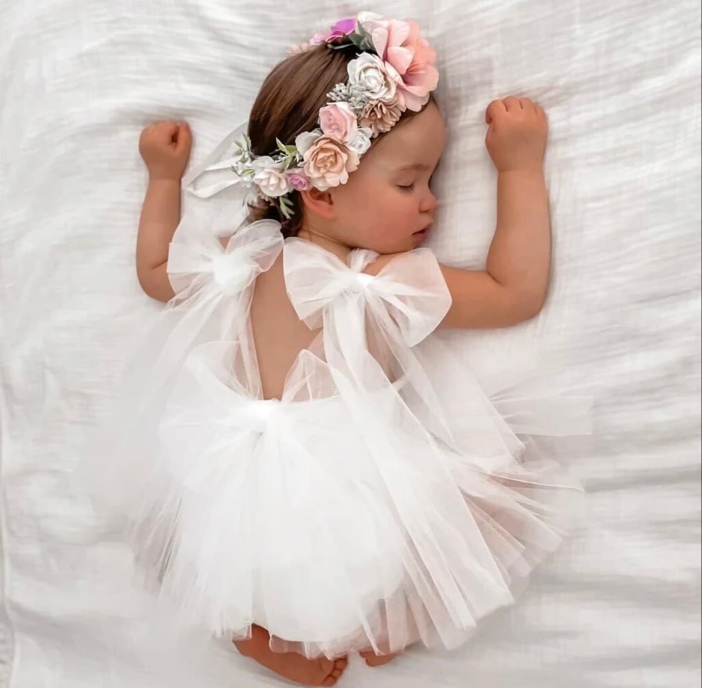 Sweet newborn baby wearing pink floral headpiece and white tulle dress sleeping peacefully on white blanket.