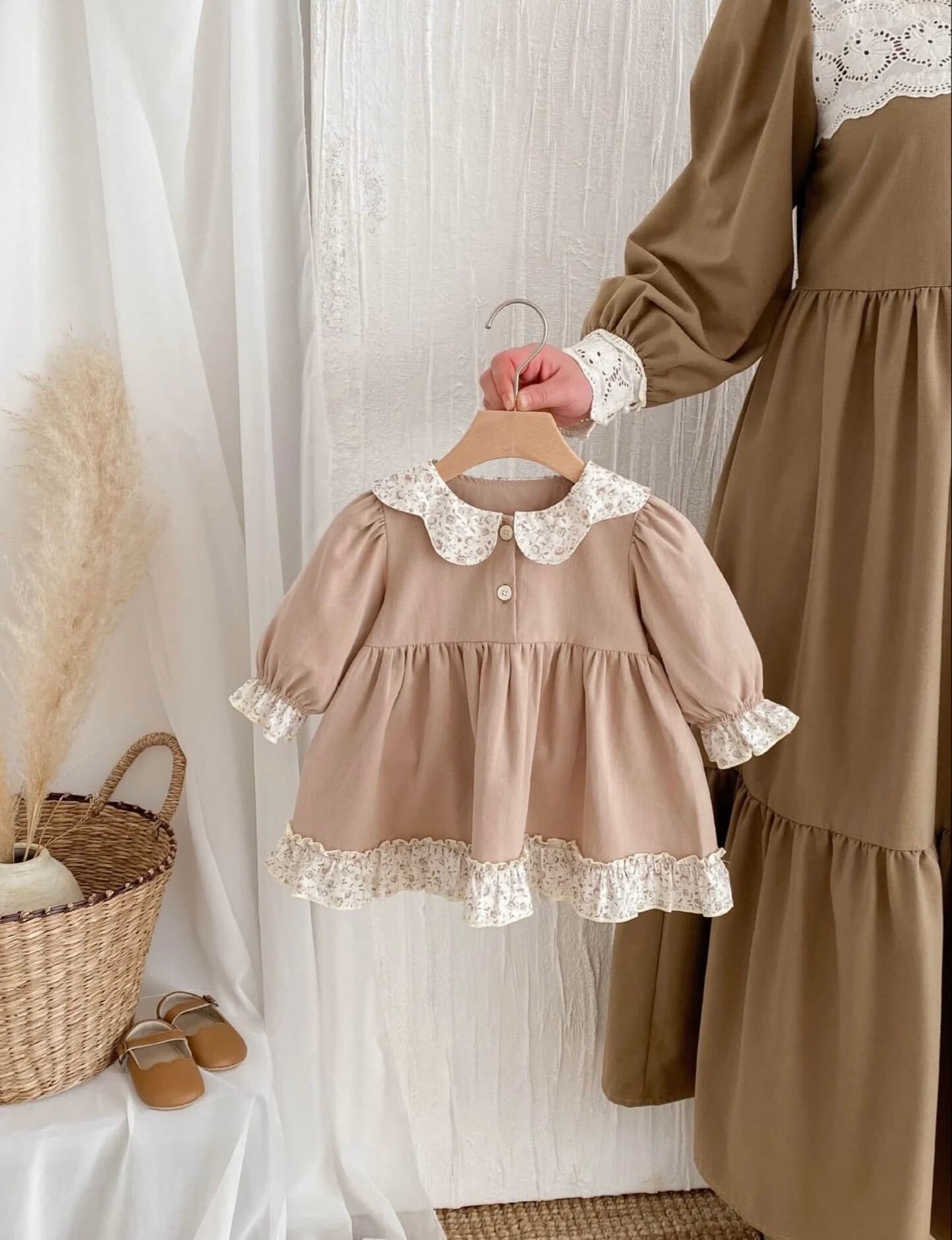 A vintage-style beige toddler dress with white lace collar and trim hangs on display next to a matching adult dress.