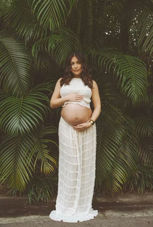A pregnant woman in a white lace dress poses against lush tropical palm fronds in an outdoor setting.