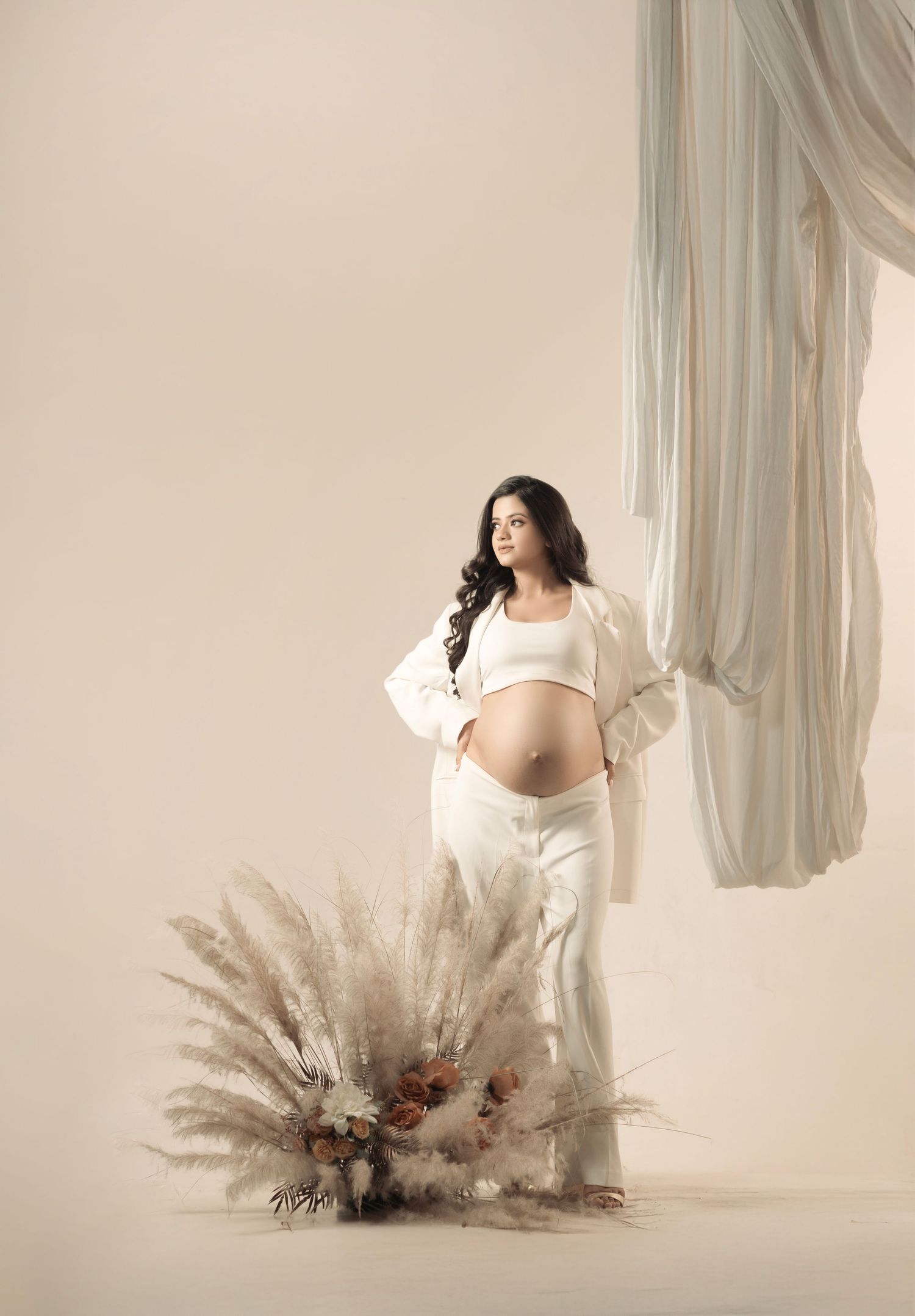 Artistic maternity photography with pampas grass arrangement and flowing white fabric.