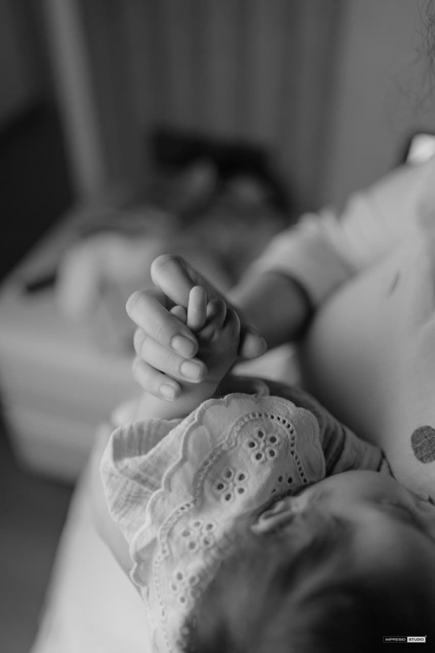 Black and white photo of a newborn baby grasping an adult finger, wearing lace clothing, with a blurred parent in background.