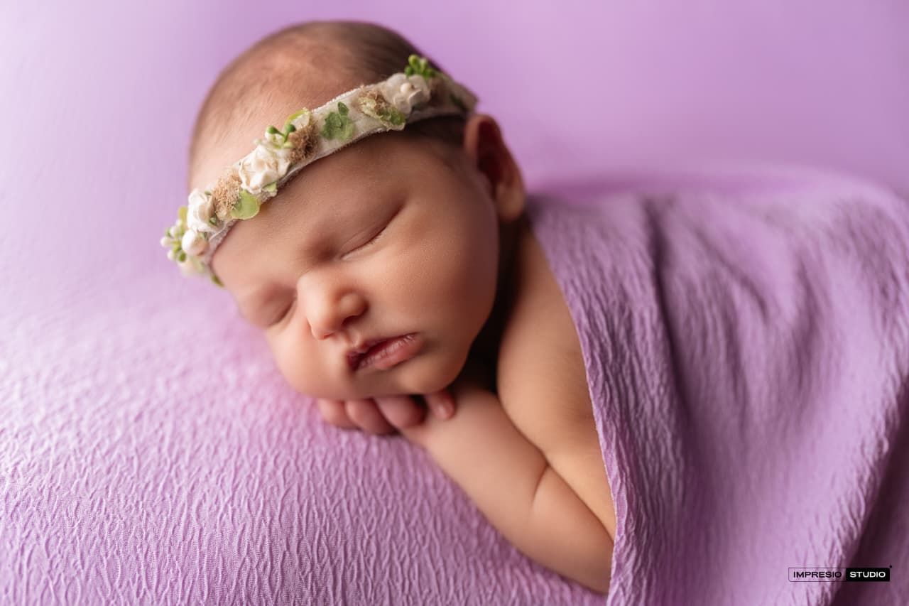 Newborn baby girl sleeping peacefully wrapped in lavender with a delicate floral headband.