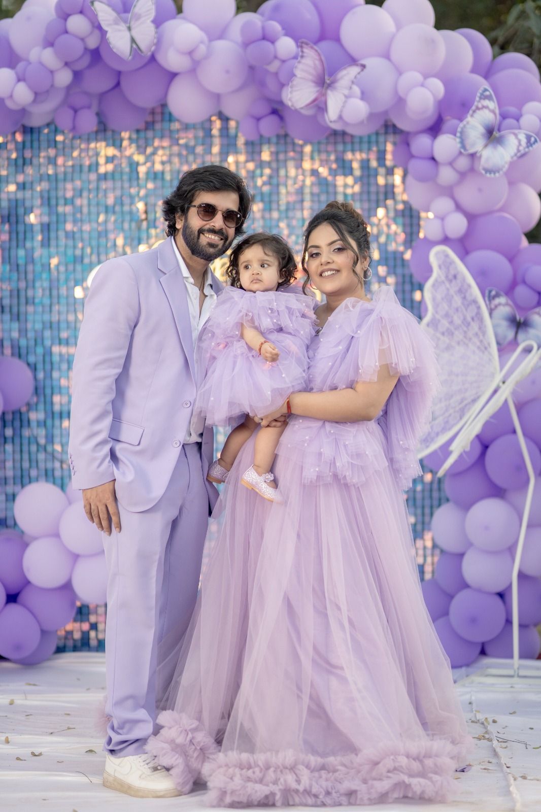 Family in matching lavender outfits posing at a purple balloon arch birthday celebration with butterfly decorations.