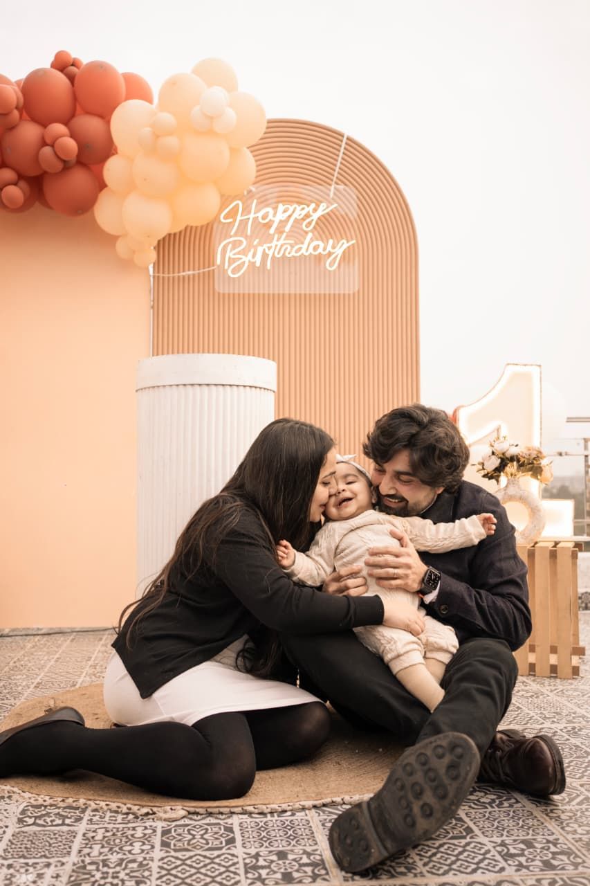 Family celebrating baby’s first birthday, parents kissing child in front of Happy Birthday sign and balloon arch.