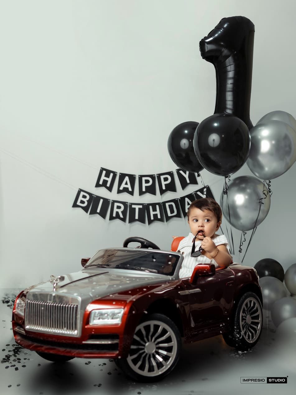 Baby sits in a toy Rolls-Royce car at a first birthday party with black and silver balloons and Happy Birthday banner.