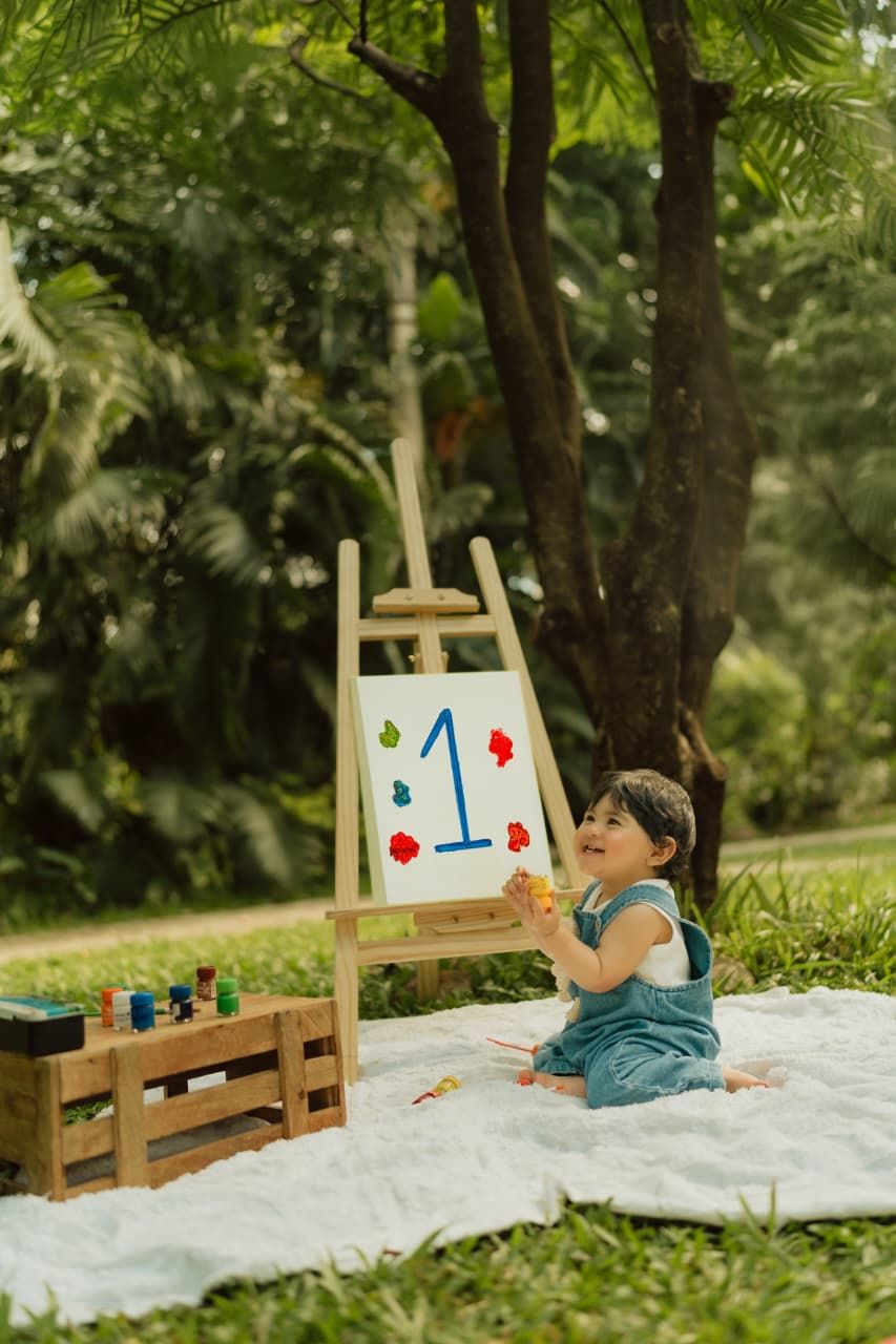 Toddler in denim overalls sits on blanket outdoors beside a painted easel displaying number 1 for first birthday.