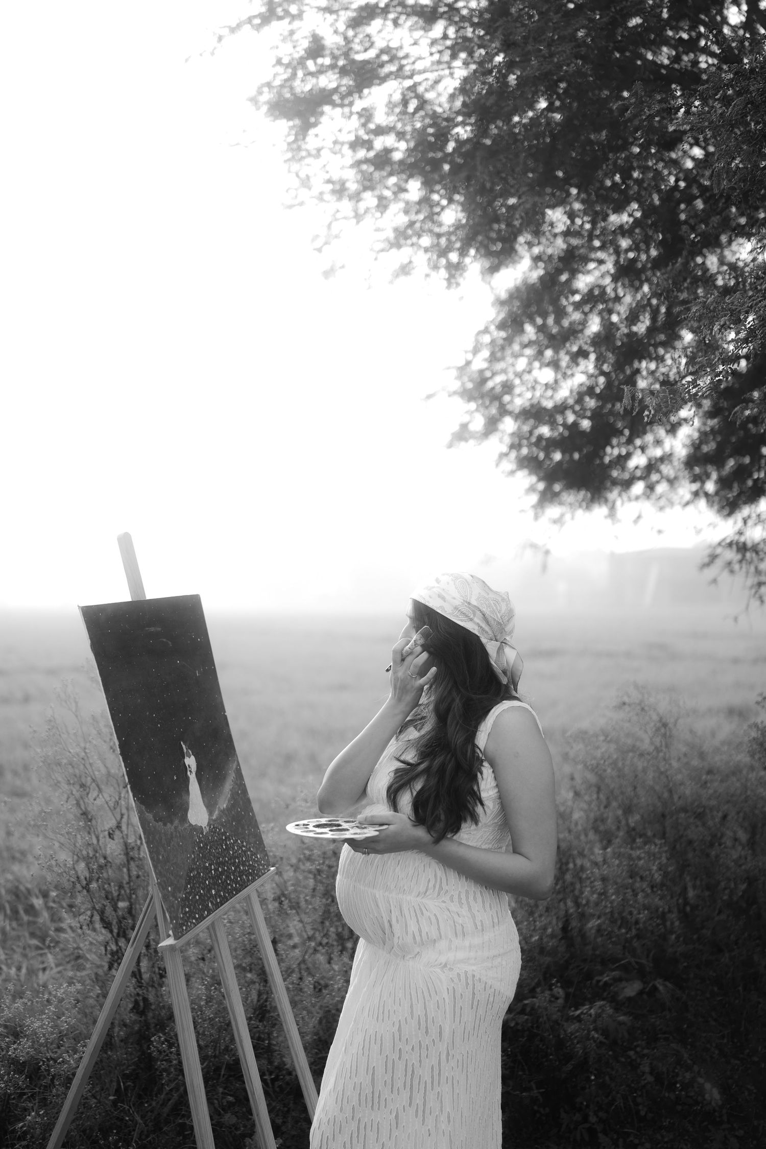 Black and white photo sequence shows an artist in a light dress painting at an easel outdoors in a misty field.