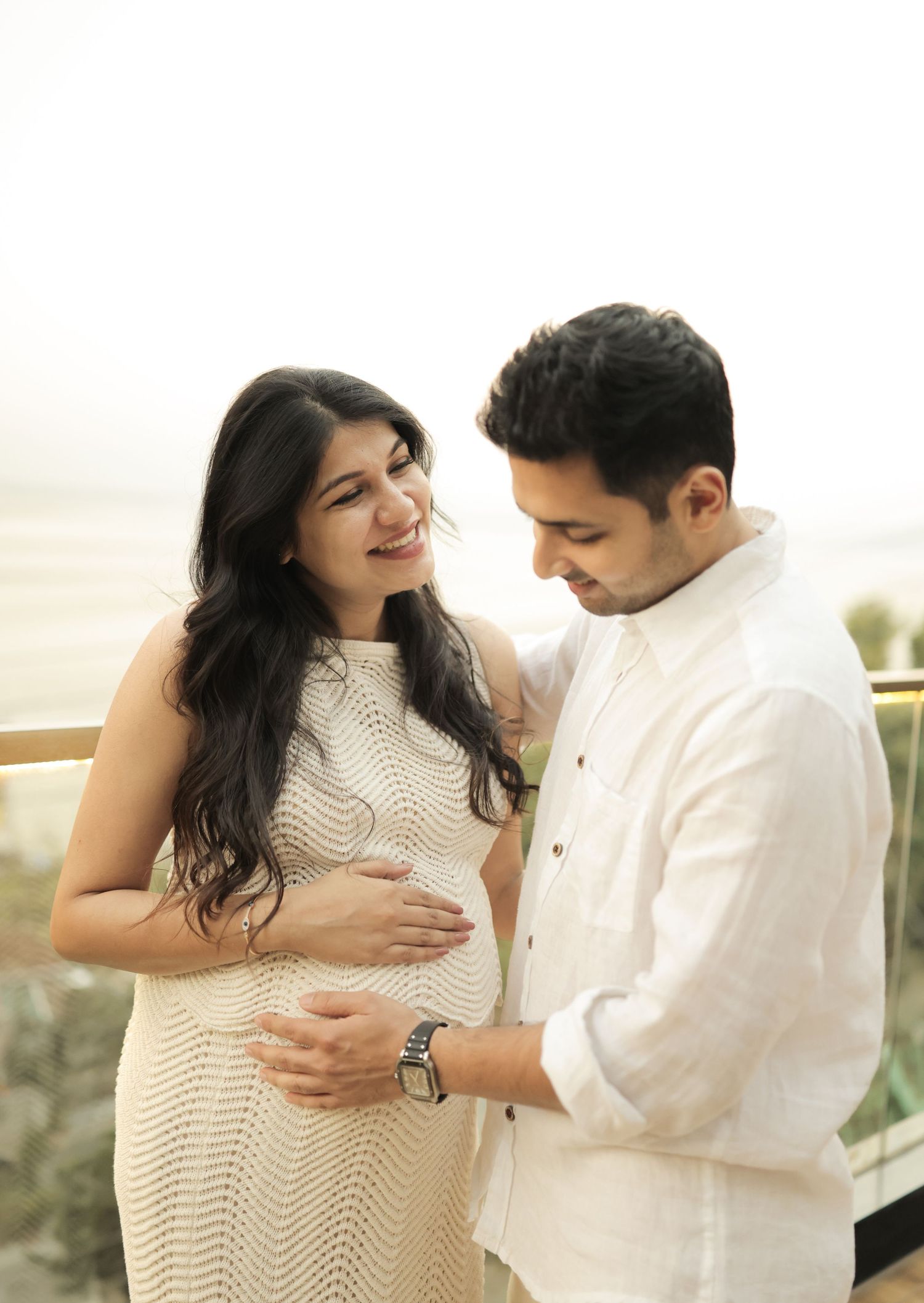 Happy expectant couple in white outfits sharing tender moment on balcony during maternity photoshoot.