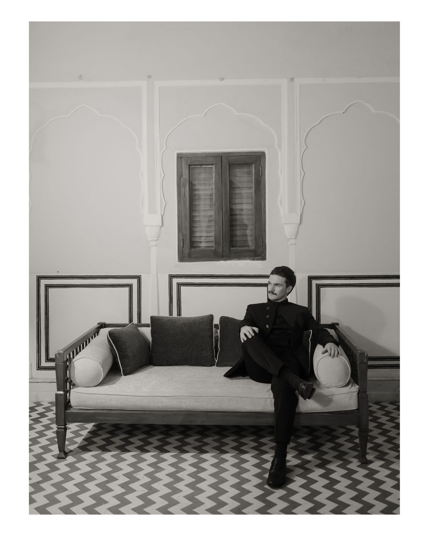 Black and white photo of a man in dark suit sitting on an elegant sofa in a decorated room with arched wall details.