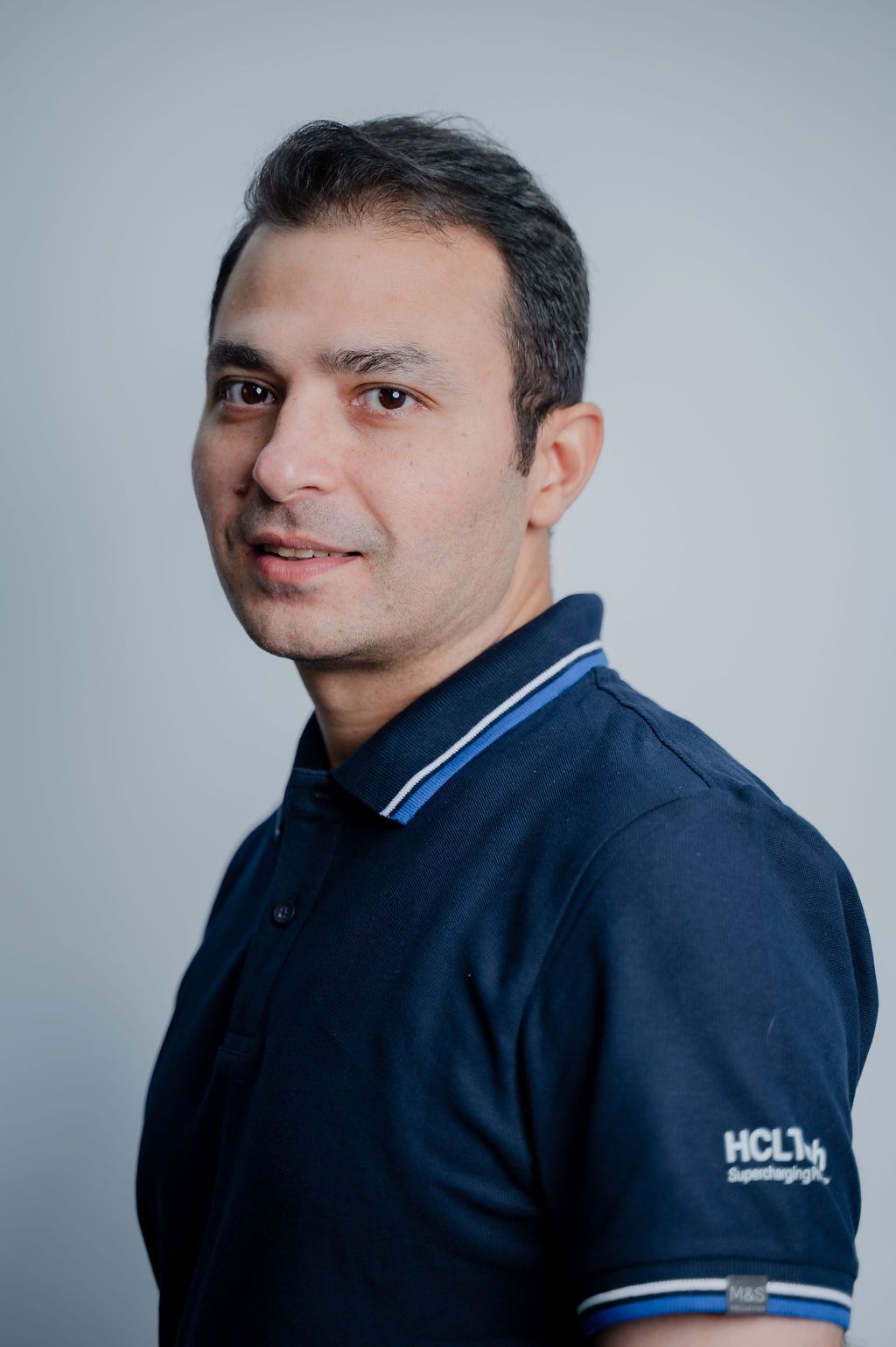 A professional man in a navy HCL polo shirt poses for a corporate headshot against a light gray background.