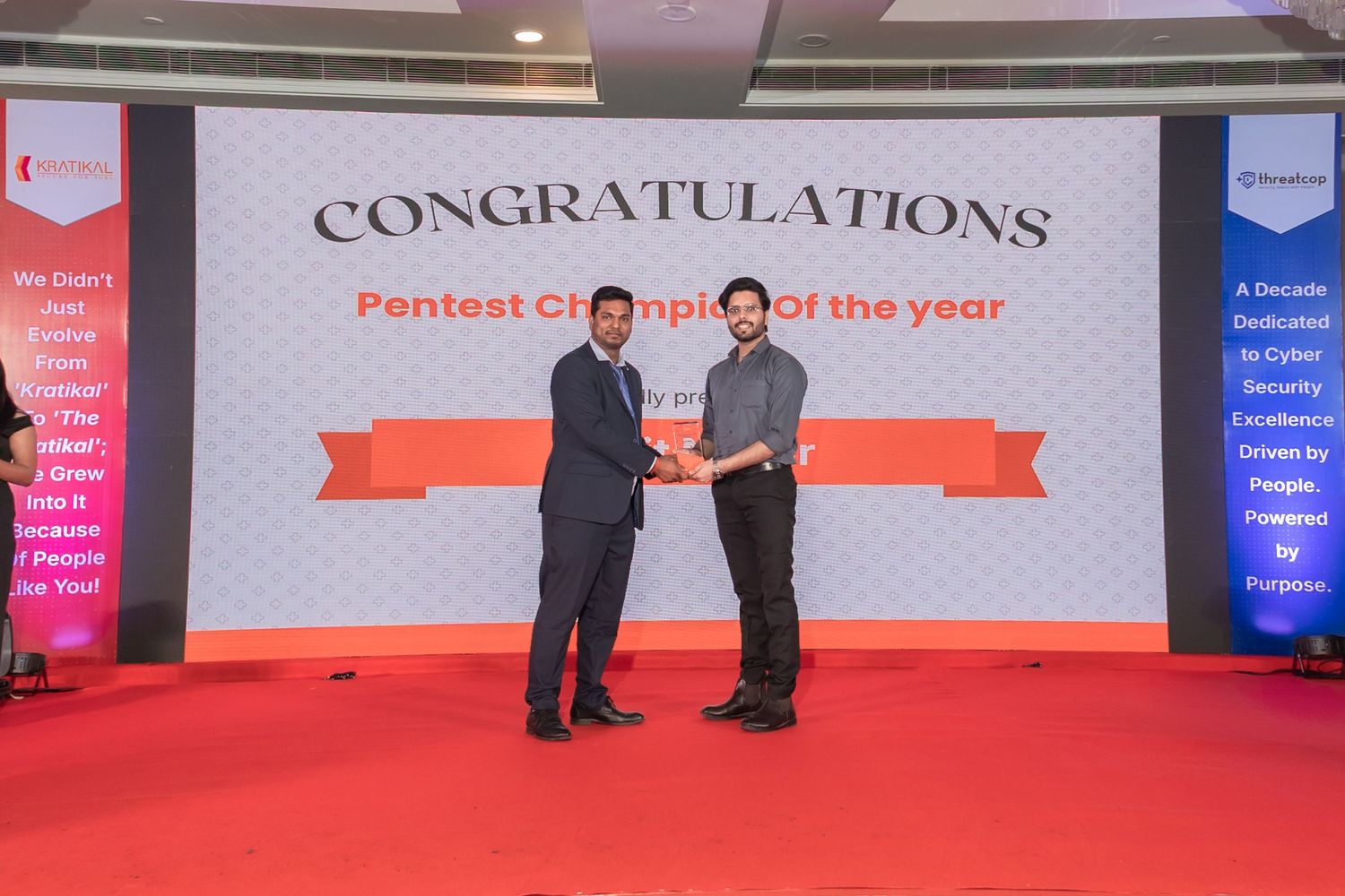 Two men in formal attire shake hands on a red stage during a Pentest Champion of the Year award ceremony with a congratulations banner.