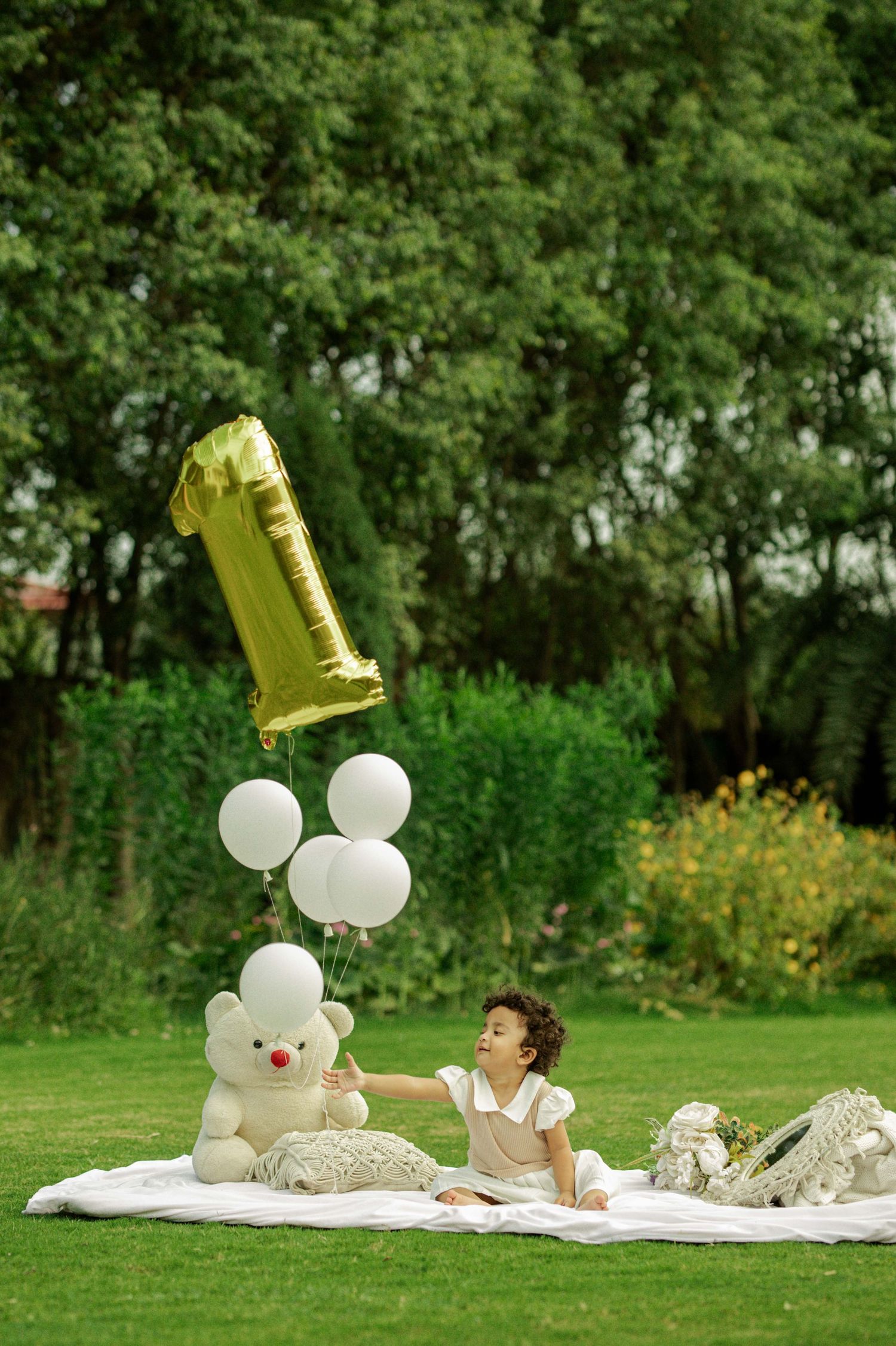 A baby girl celebrates her 1st birthday on a white blanket in a garden with a teddy bear and gold number one balloon.