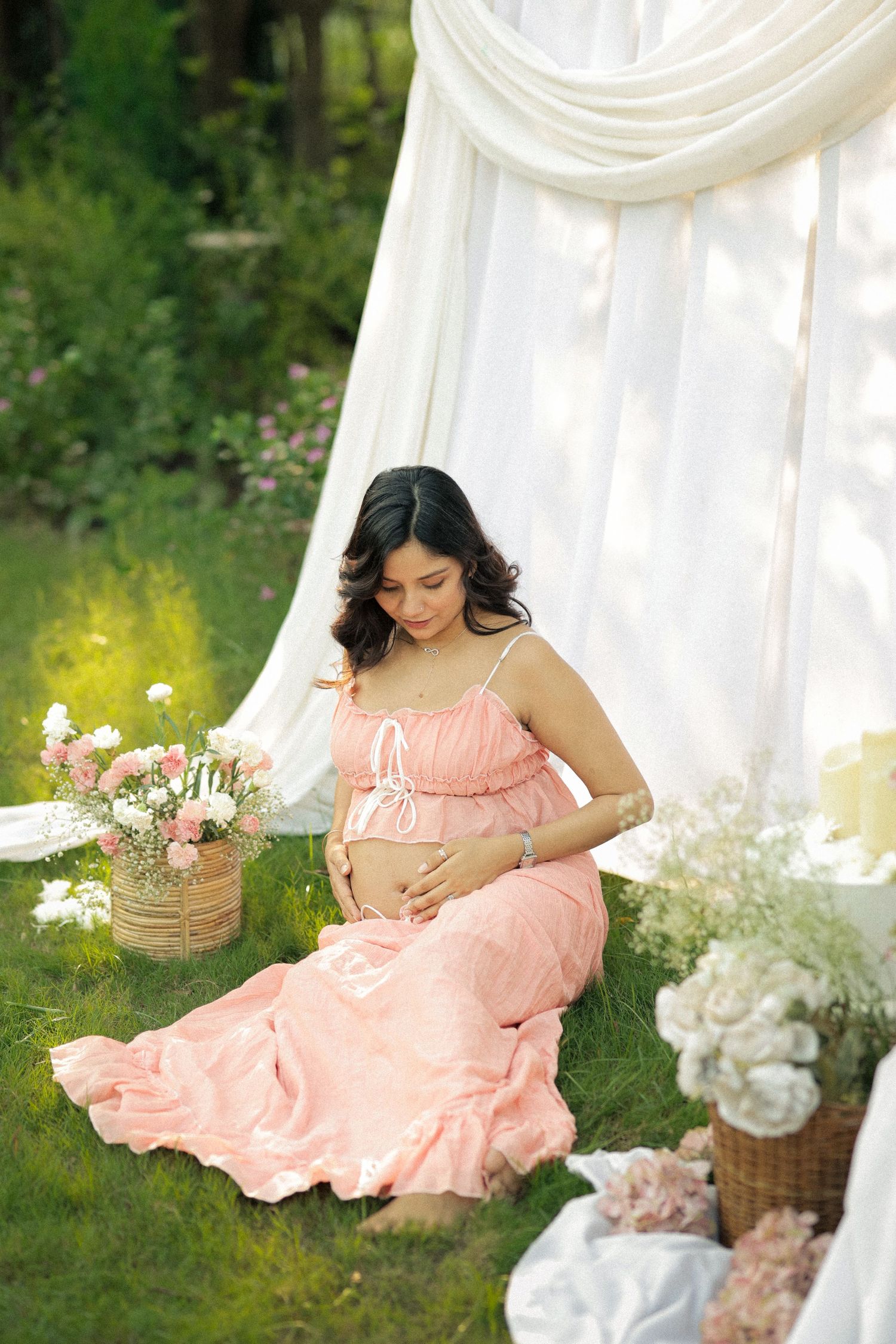 Pregnant woman in pink dress sitting on grass near white drapes and flowers, cradling her baby bump outdoors.