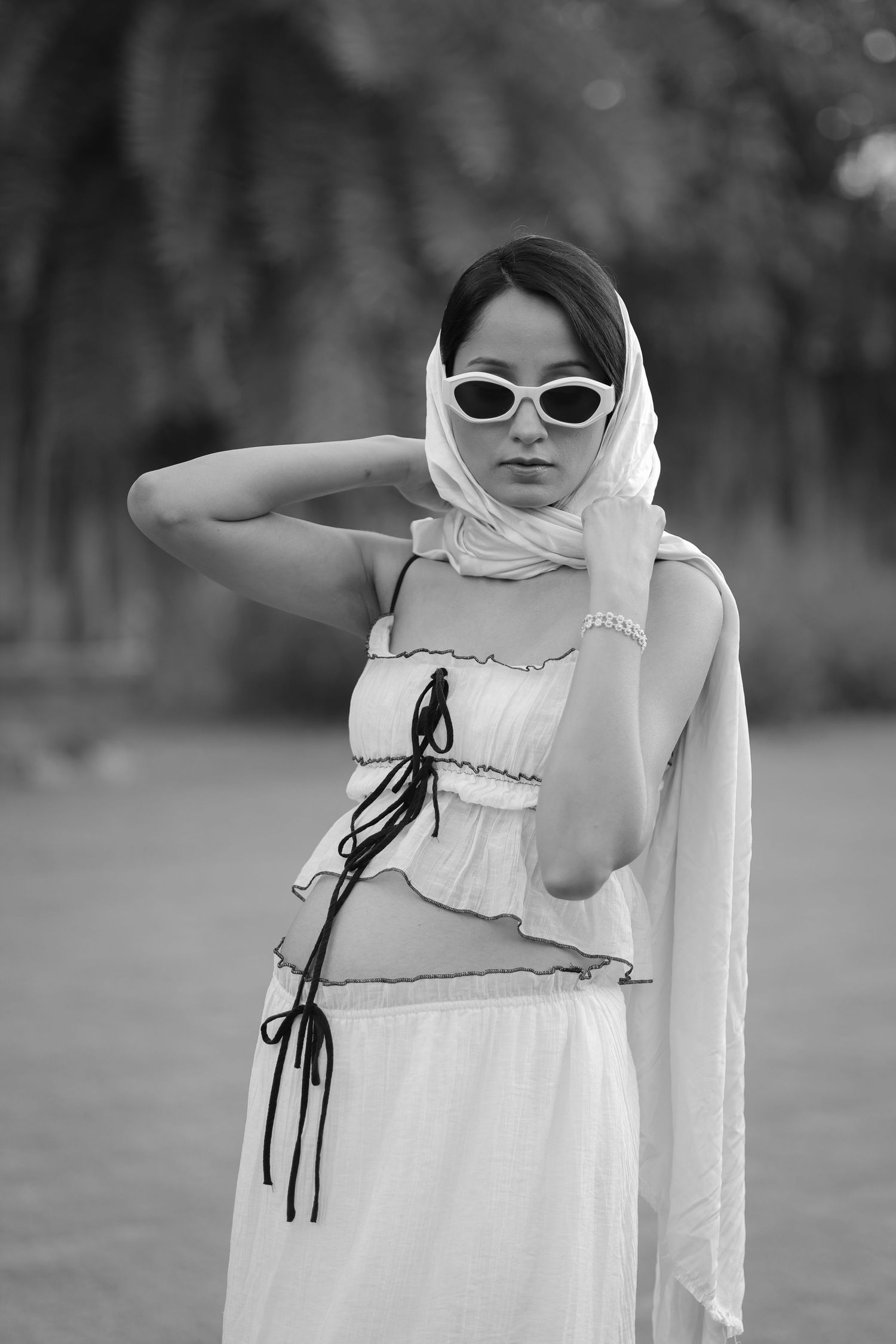 Black and white photo of a stylish woman in white outfit, head scarf, and cat-eye sunglasses posing outdoors.