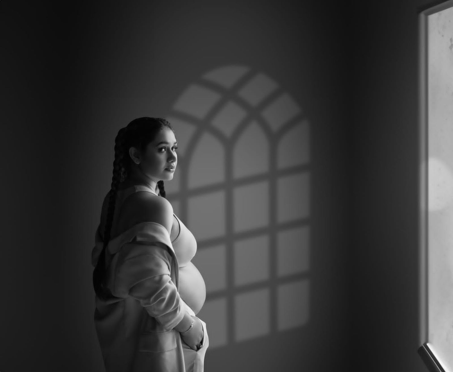 Black and white maternity photo of pregnant woman in soft robe gazing at arched window with dramatic shadows.
