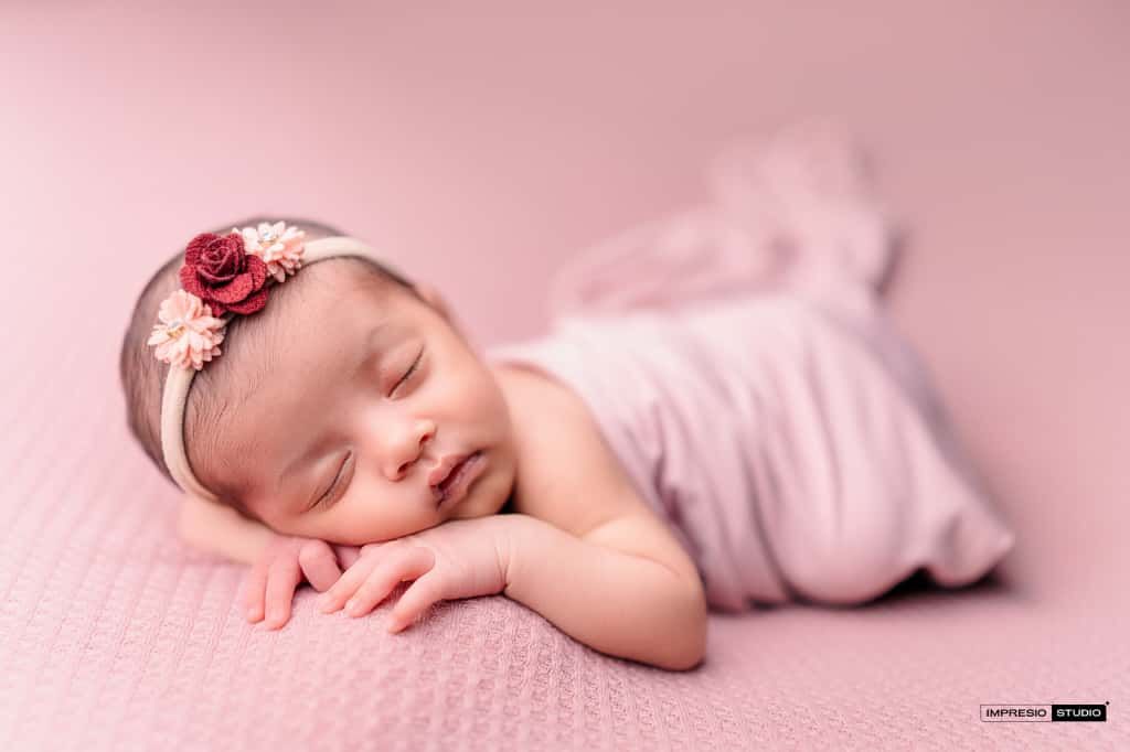 Sleeping newborn baby girl in pink wrap and floral headband on pink background.
