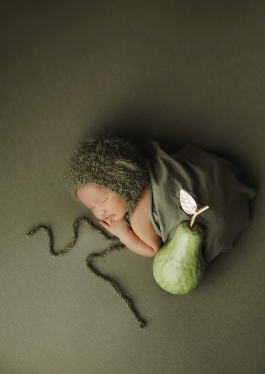 Newborn baby wrapped in green fabric with a fuzzy hat, posed beside a fresh pear on a green background.
