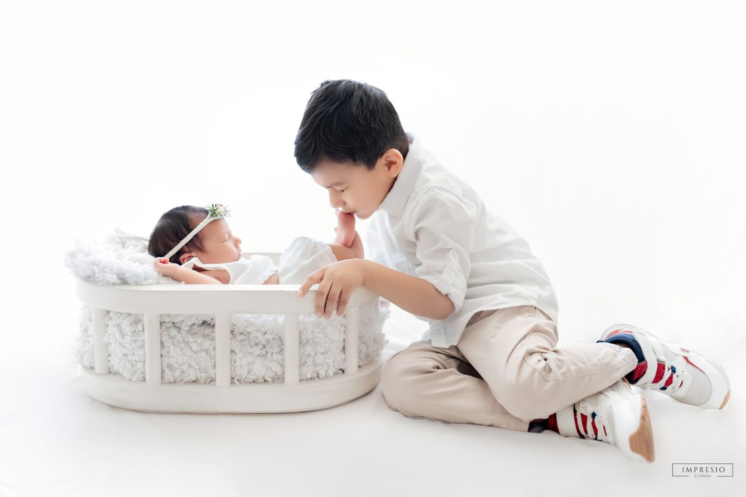 A young boy tenderly interacts with his newborn sibling resting in a white wicker bassinet during a studio photo session.