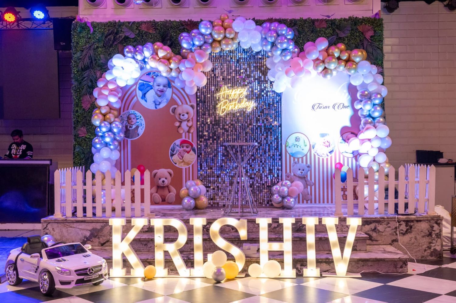 Colorful balloon arch birthday party decoration with illuminated KRISHIV letter lights and teddy bear themed backdrop.