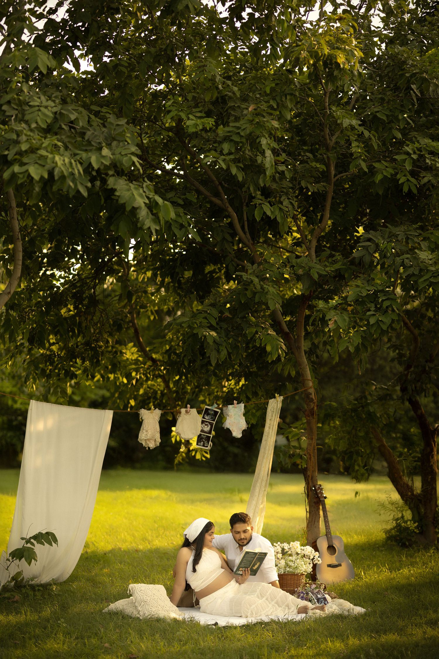 A couple relaxes on a blanket under trees with white linens hanging from clotheslines in a romantic outdoor setting.