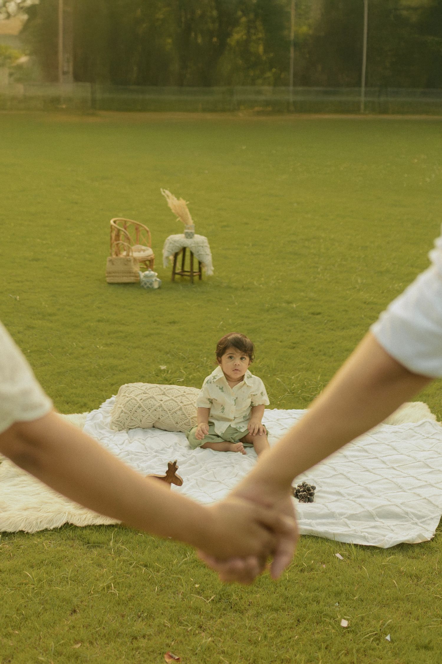 Family picnic scene on a grassy field with white blankets spread out and chairs visible in the background.