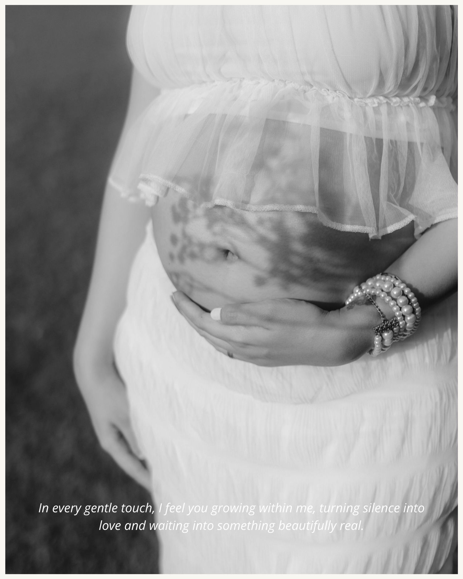 Black and white maternity photo of pregnant woman in white dress cradling baby bump with pearl bracelets.