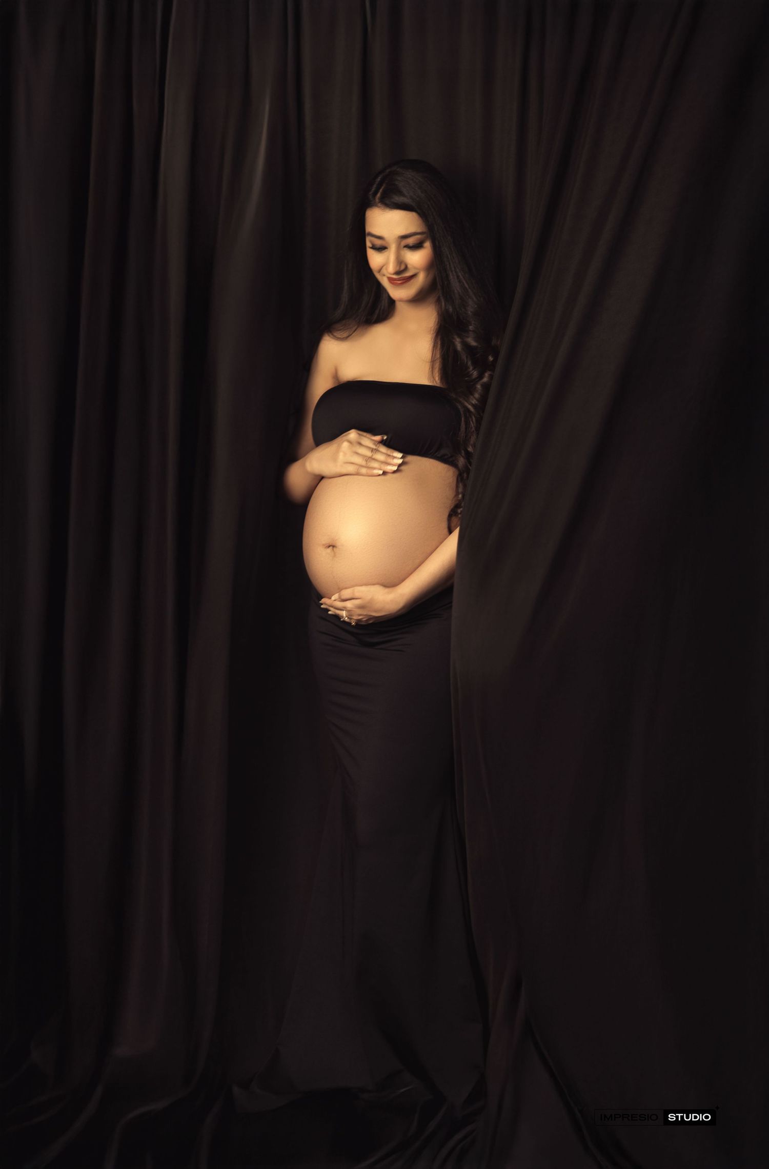 Elegant pregnant woman in black off-shoulder gown cradling baby bump against dark draped backdrop.