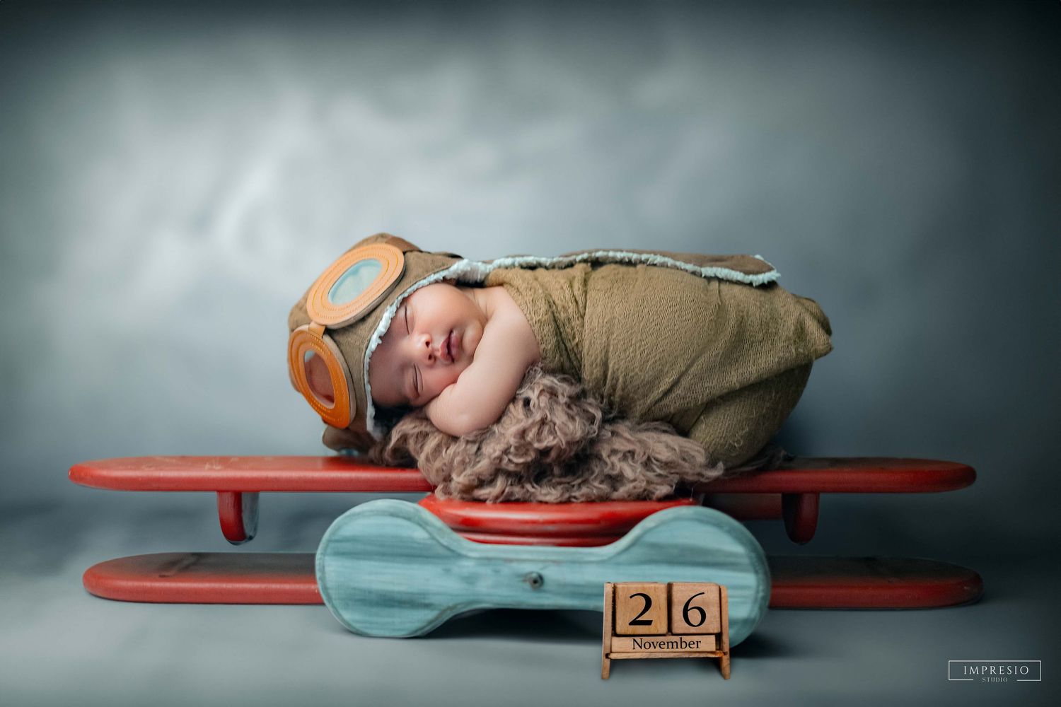 Newborn baby sleeping on red toy airplane wearing aviator hat with number 26 blocks in soft studio photography.