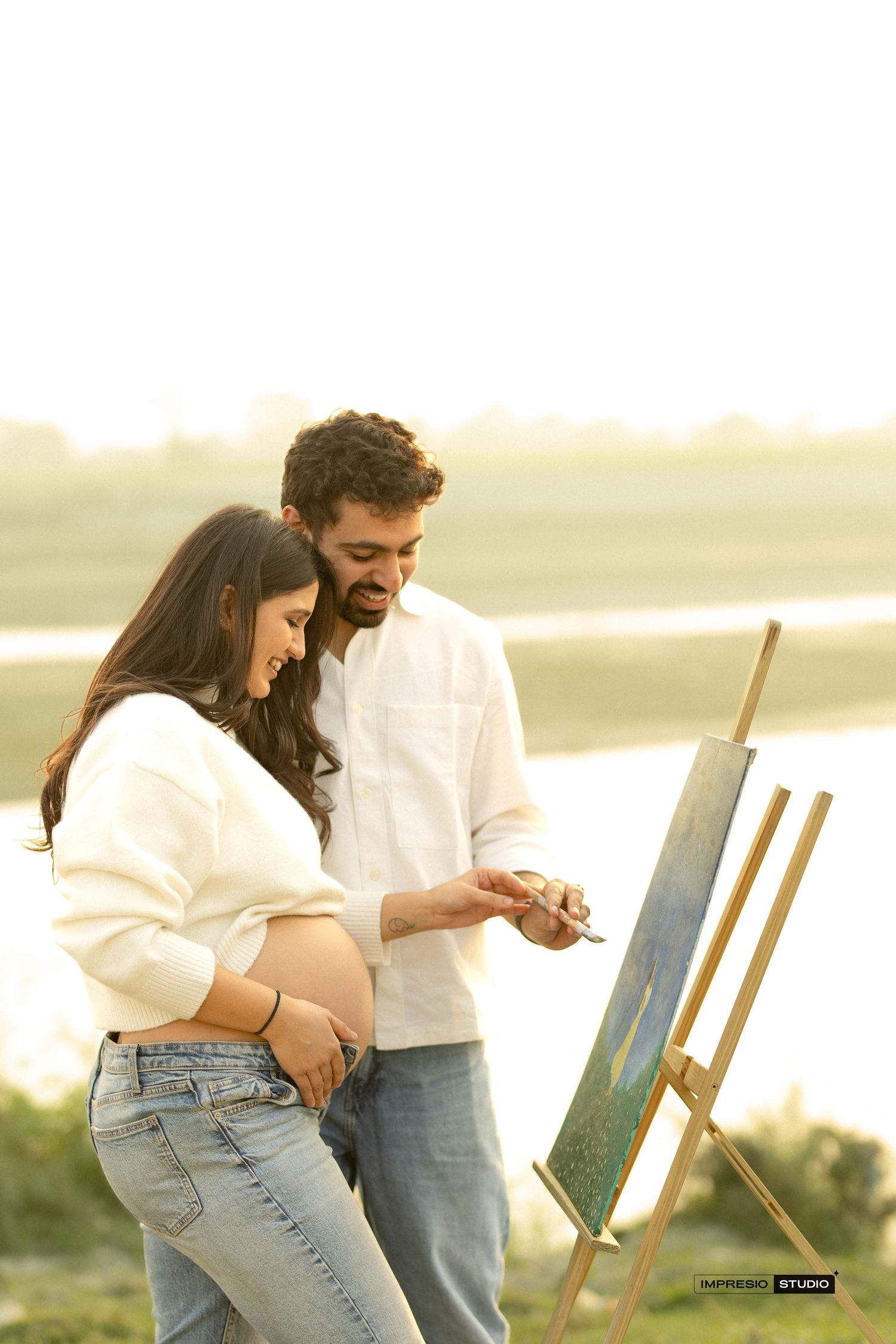 Pregnant couple painting on canvas outdoors at golden hour, smiling together near a serene waterfront.