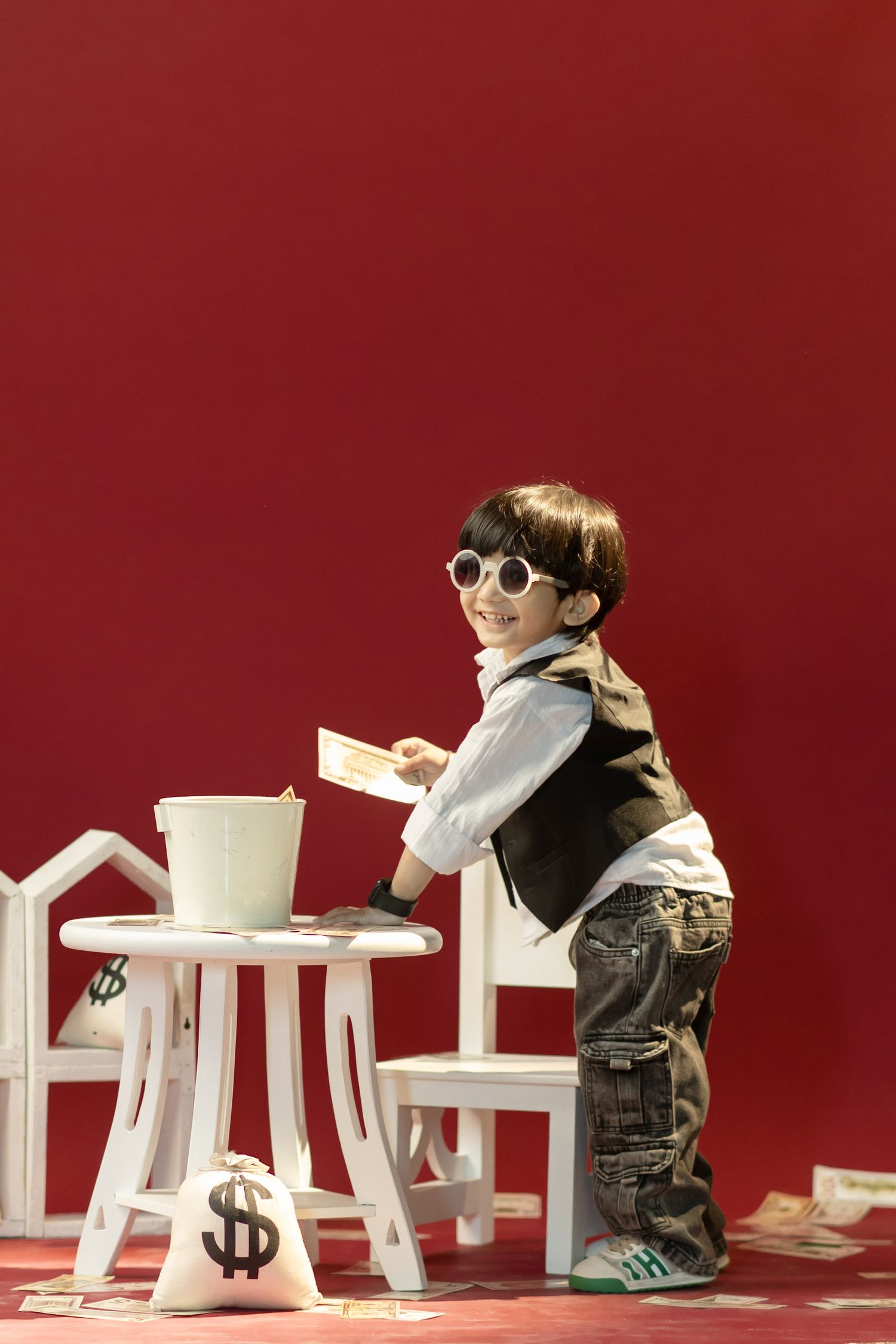 Young boy in sunglasses and vest playing with toy houses and money bags on white table against red background.