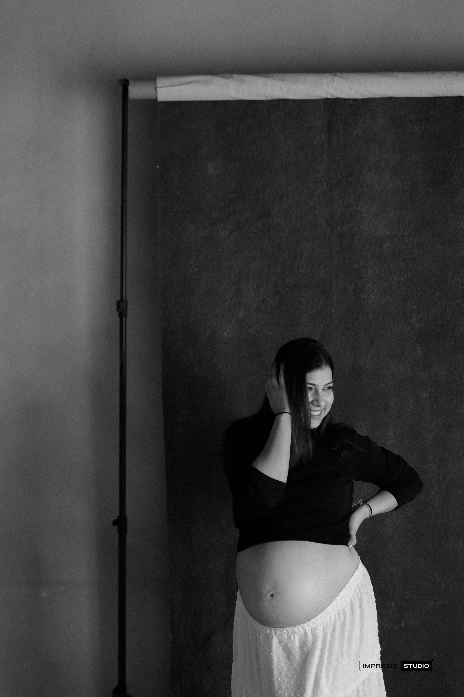 Black and white maternity photo of pregnant woman in black top and white skirt posing in photography studio.