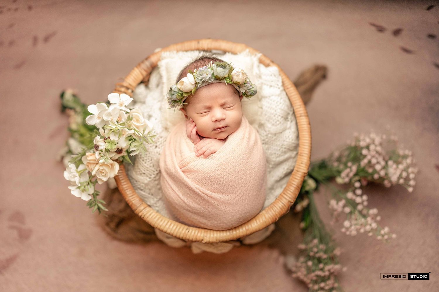 Sleeping newborn wrapped in peach swaddle, nestled in a basket with a floral crown and white flowers.