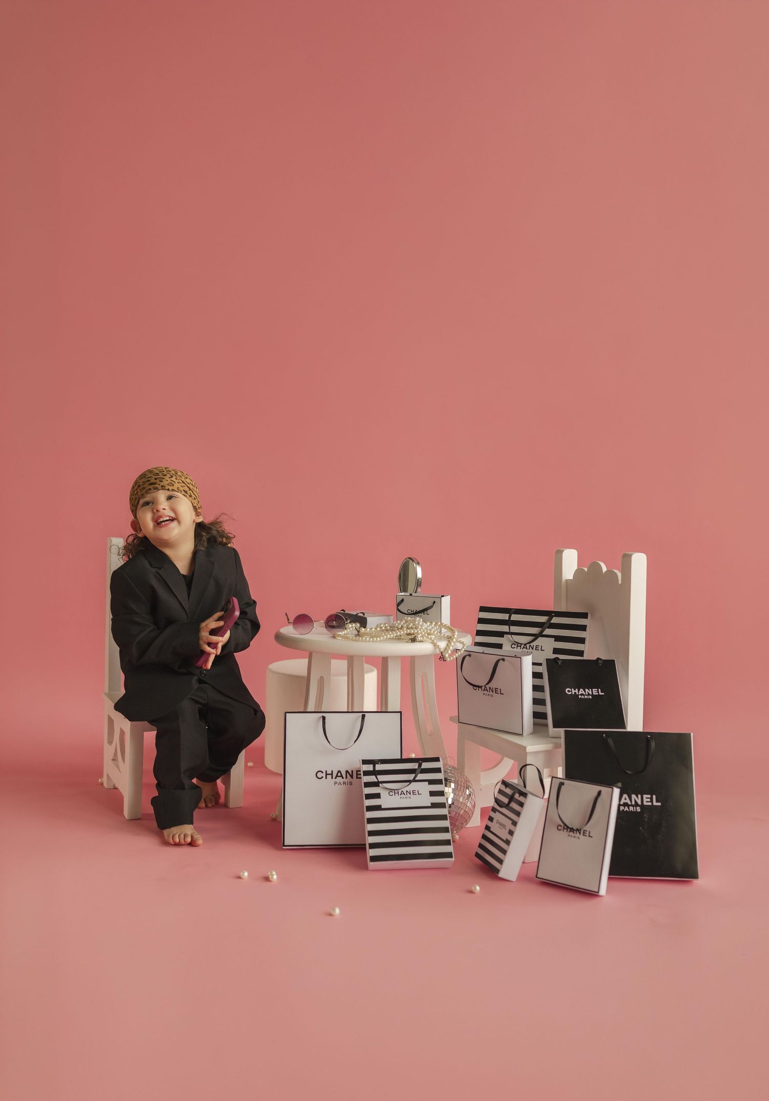 A laughing child in a black suit surrounded by Chanel shopping bags on a pink background.