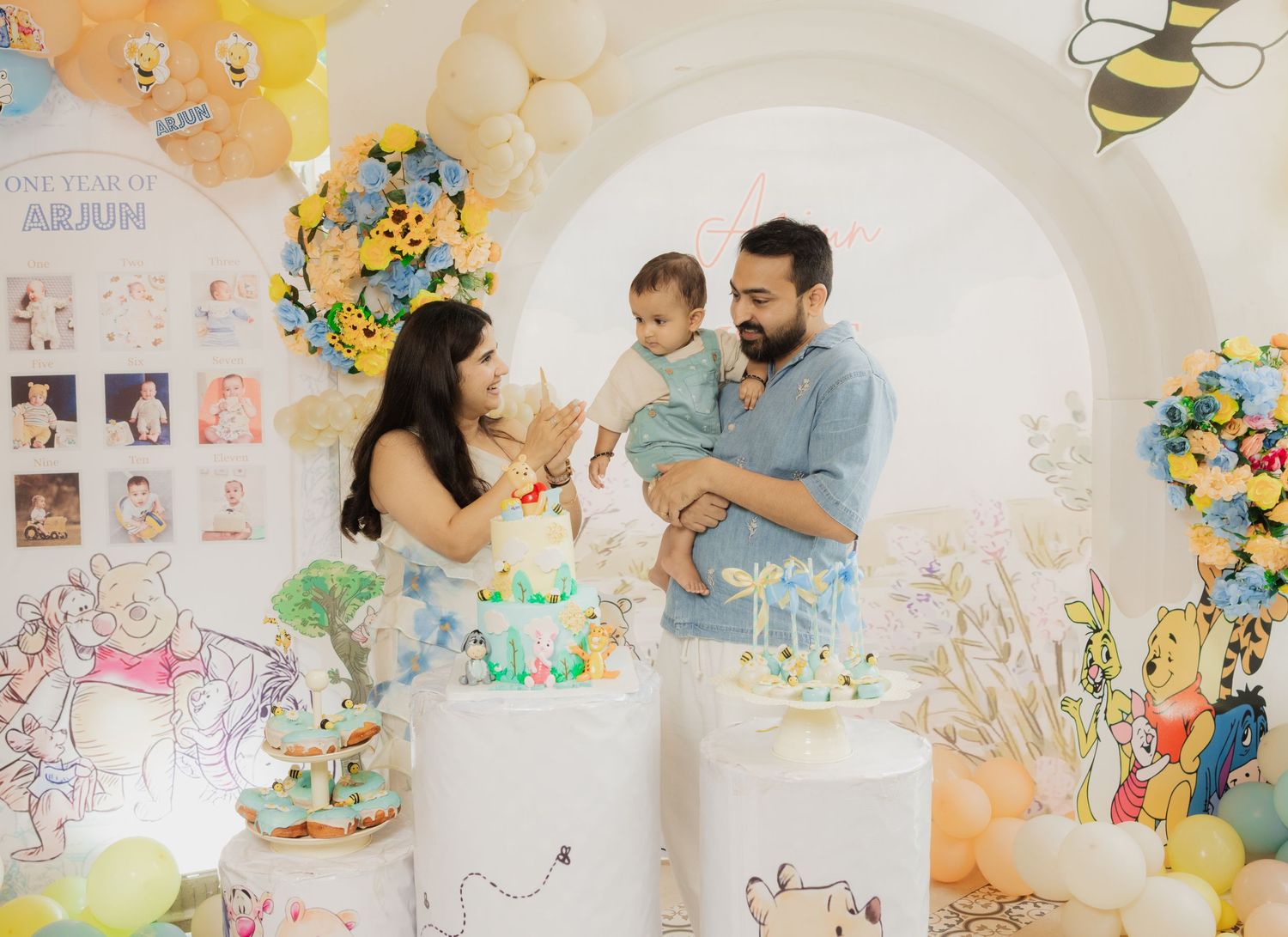 Family celebrating baby’s first birthday with Winnie the Pooh themed decor, balloons, and cake.