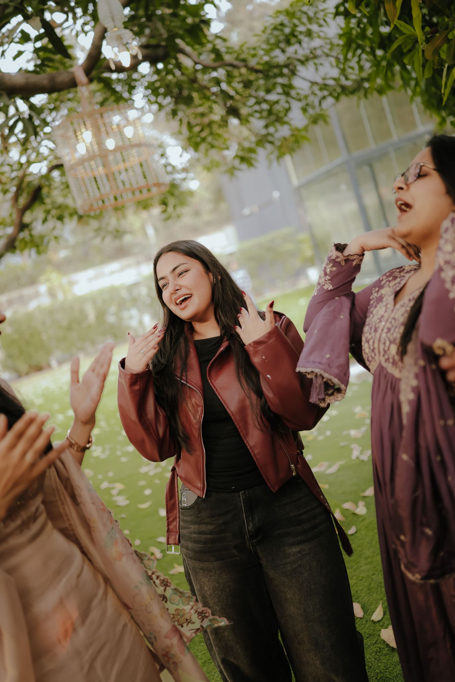 Young woman in red leather jacket laughing outdoors at garden party with friends amid falling flower petals.