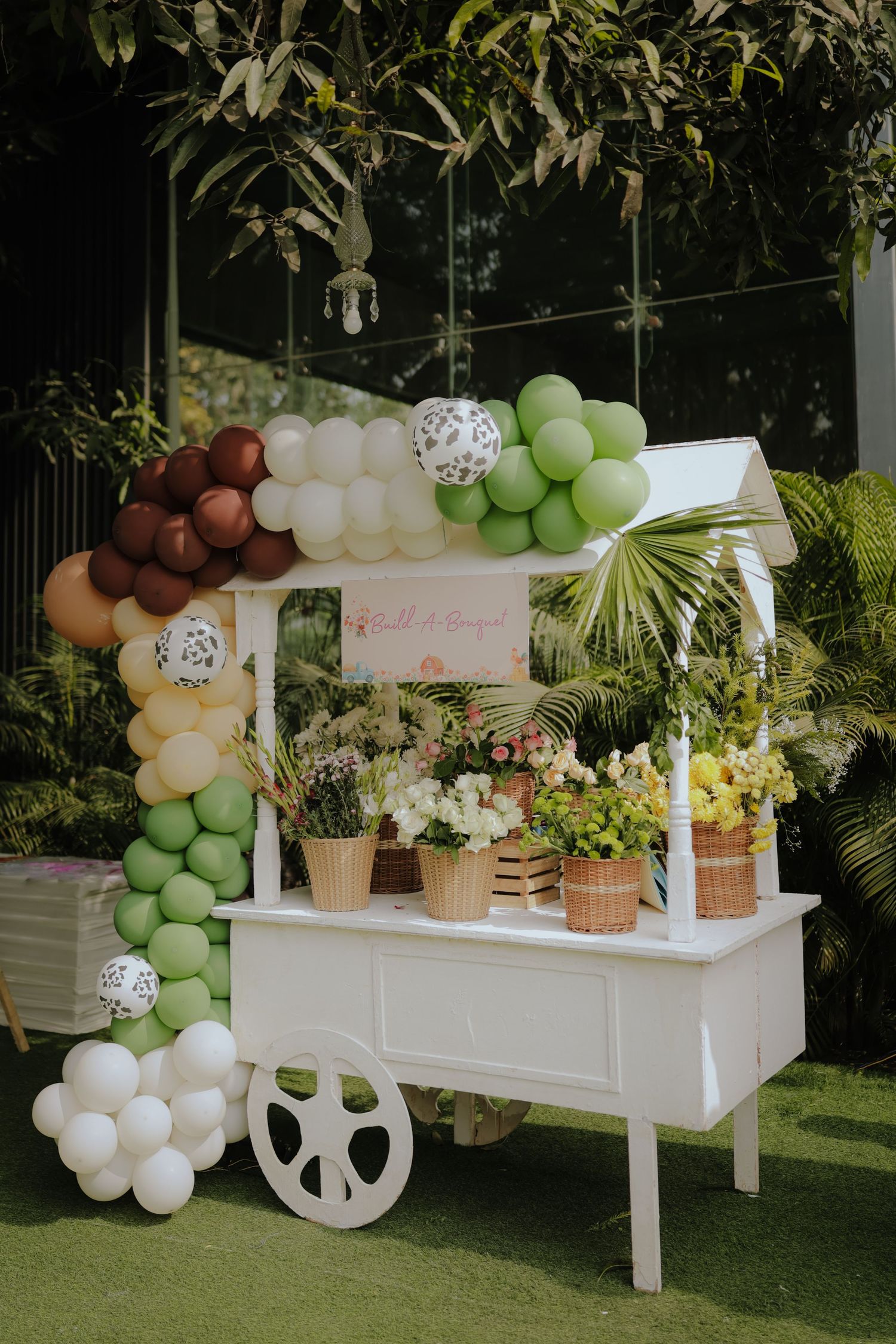 White flower cart with balloon garland in green, brown, and cream tones, surrounded by lush greenery for an outdoor party.
