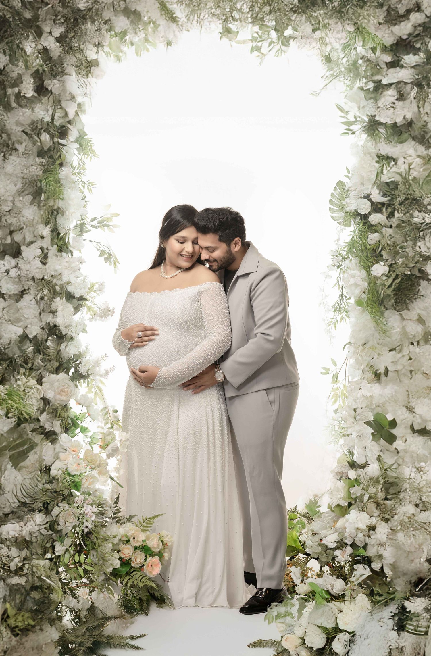 Couple in elegant maternity photoshoot under floral arch, woman in white gown cradling baby bump, man in gray suit embracing her.