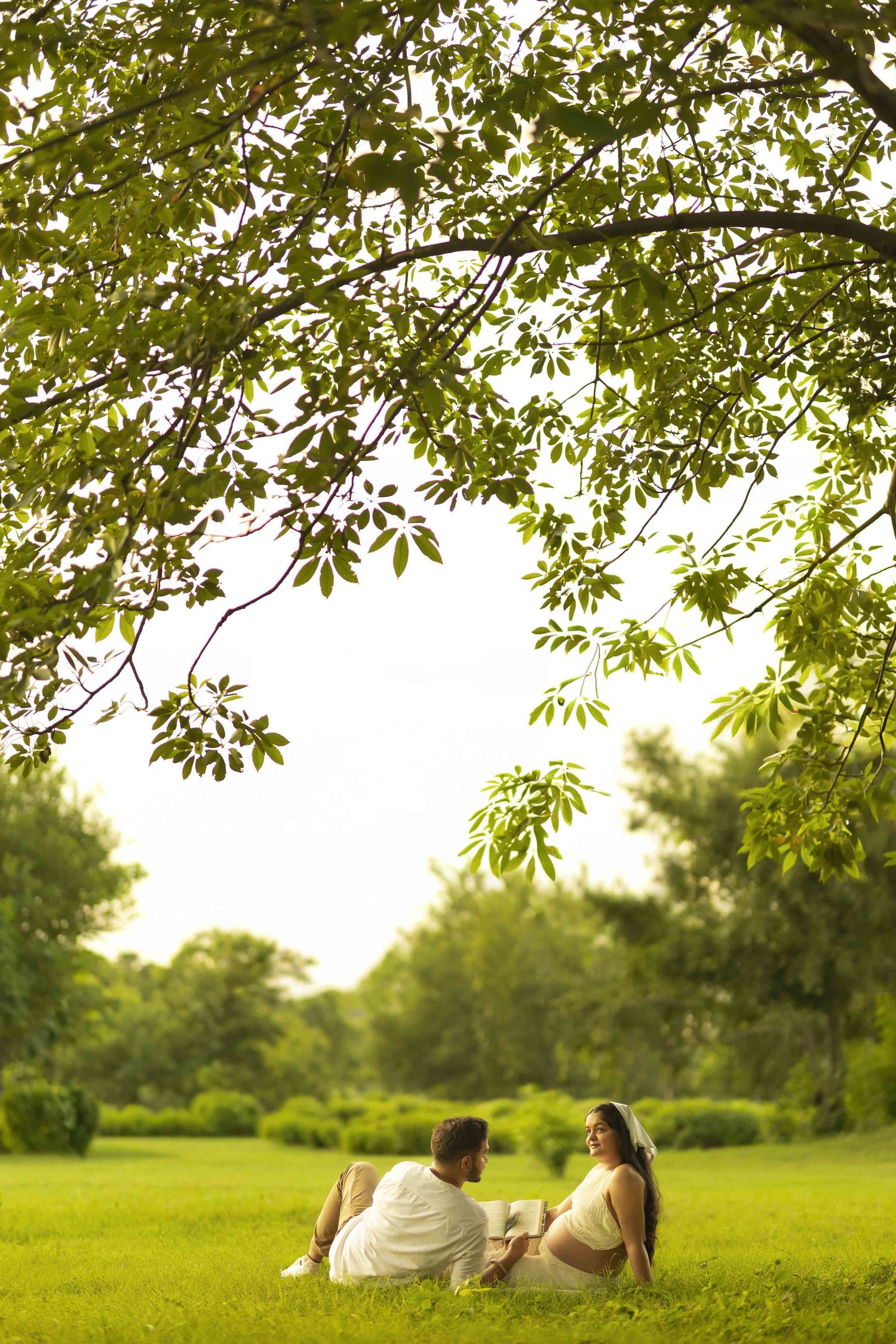 Couple sitting on green grass under a large tree in a lush park, enjoying a peaceful outdoor moment together.