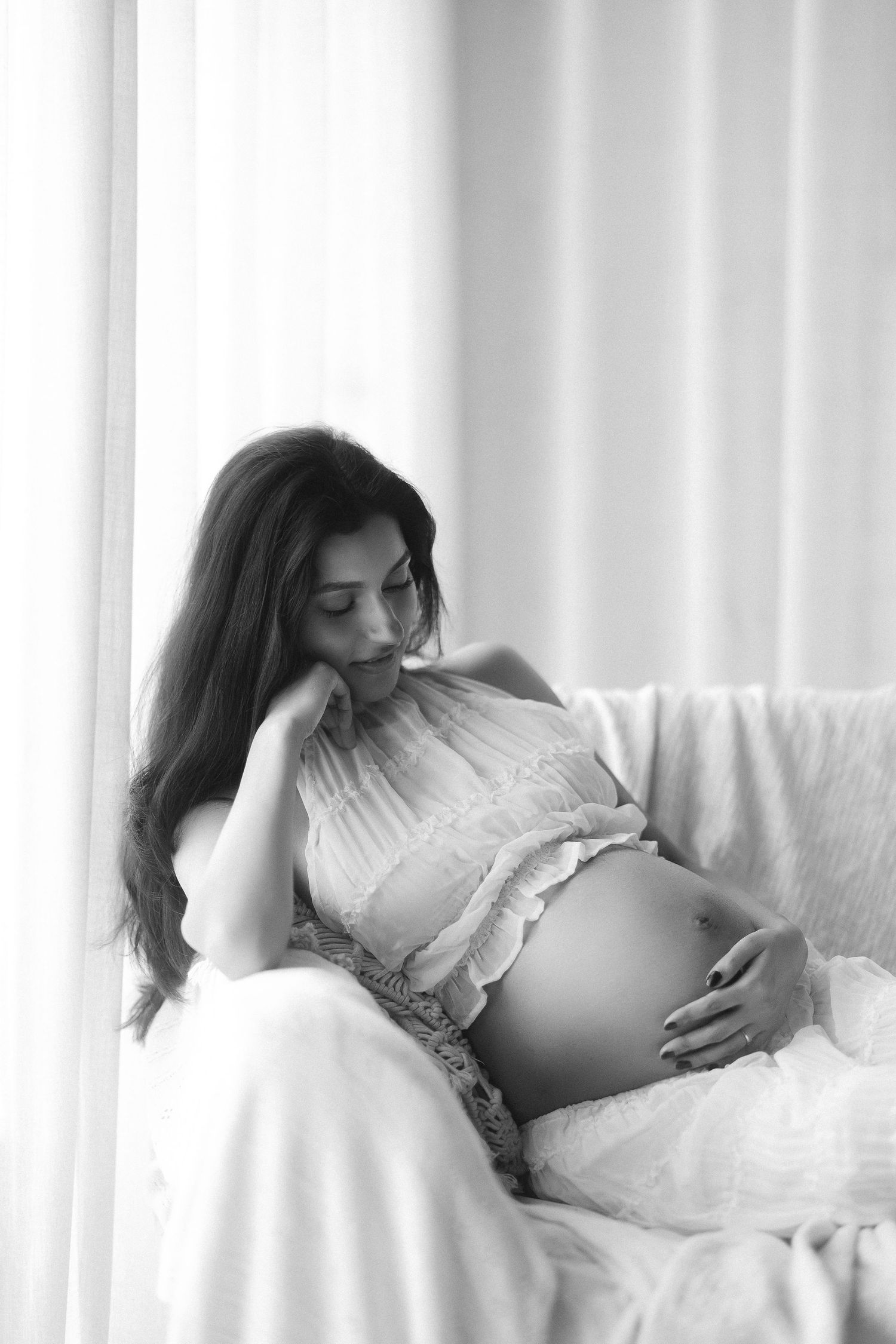 Black and white portrait of a pregnant woman sitting by a window, tenderly cradling her baby bump.